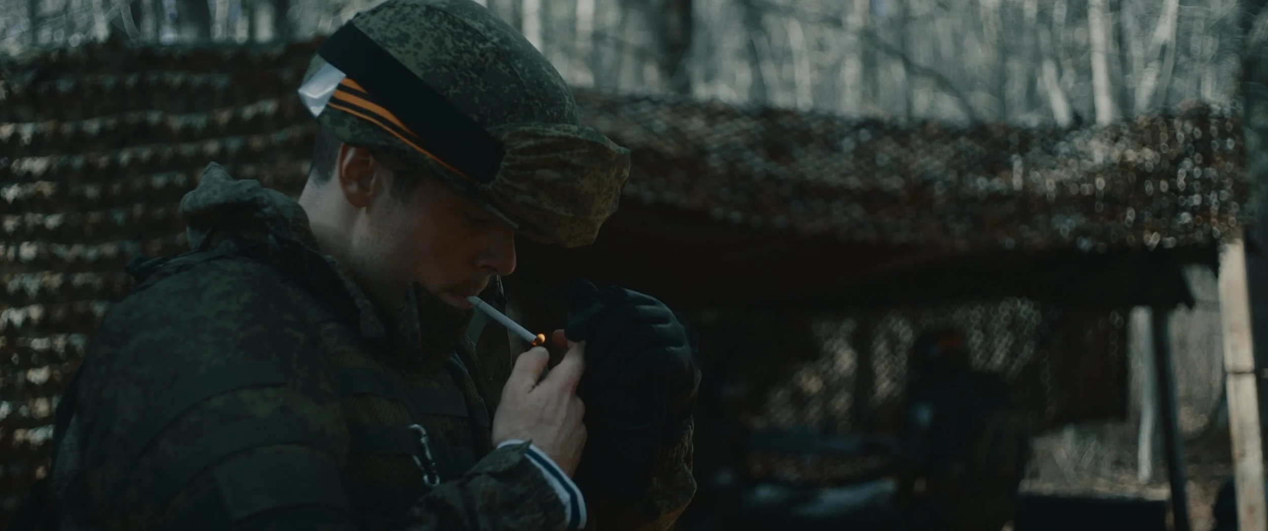 A soldier in camouflage uniform lighting a cigarette in a wooded outdoor setting.