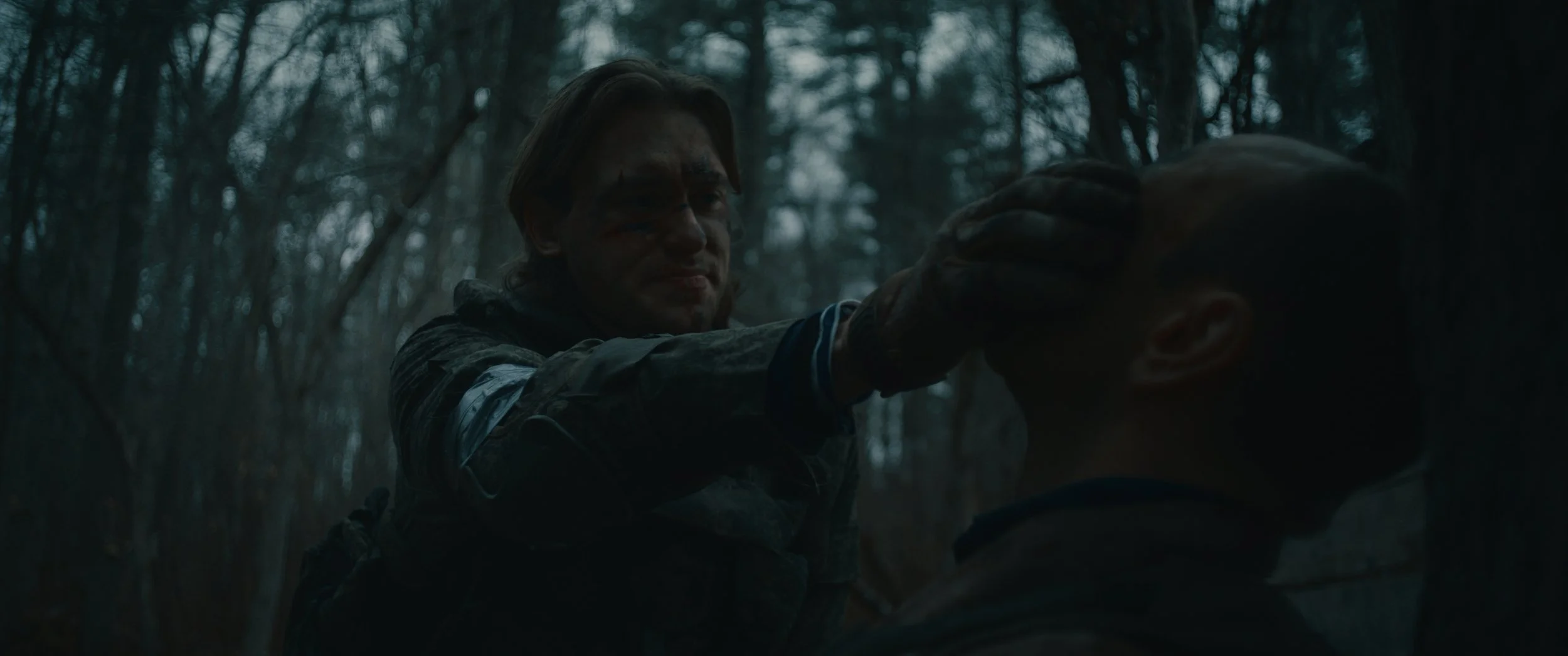 A person with long hair and a tattered face grabs another person's head in a dark, dense forest.