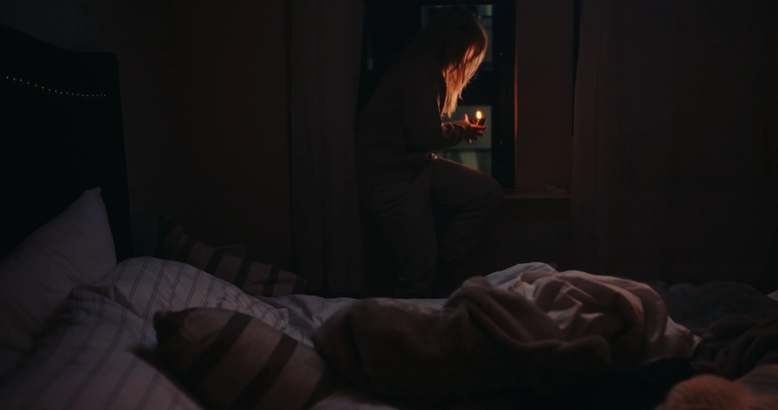 A person sitting on a windowsill holding a lit candle in a dimly lit room.