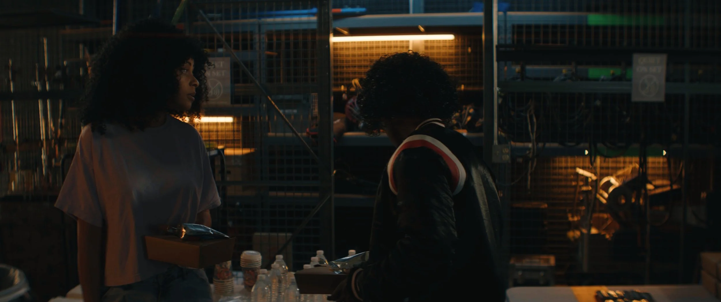 Two women are at a table in a hardware store, one receiving a box from the other. The store has metal shelves with power tools and supplies, and bottles of water on the table, with warm lighting.