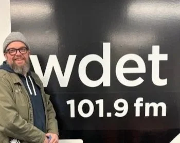 Man in glasses standing in front of a black sign that reads "WDET 101.9 FM"