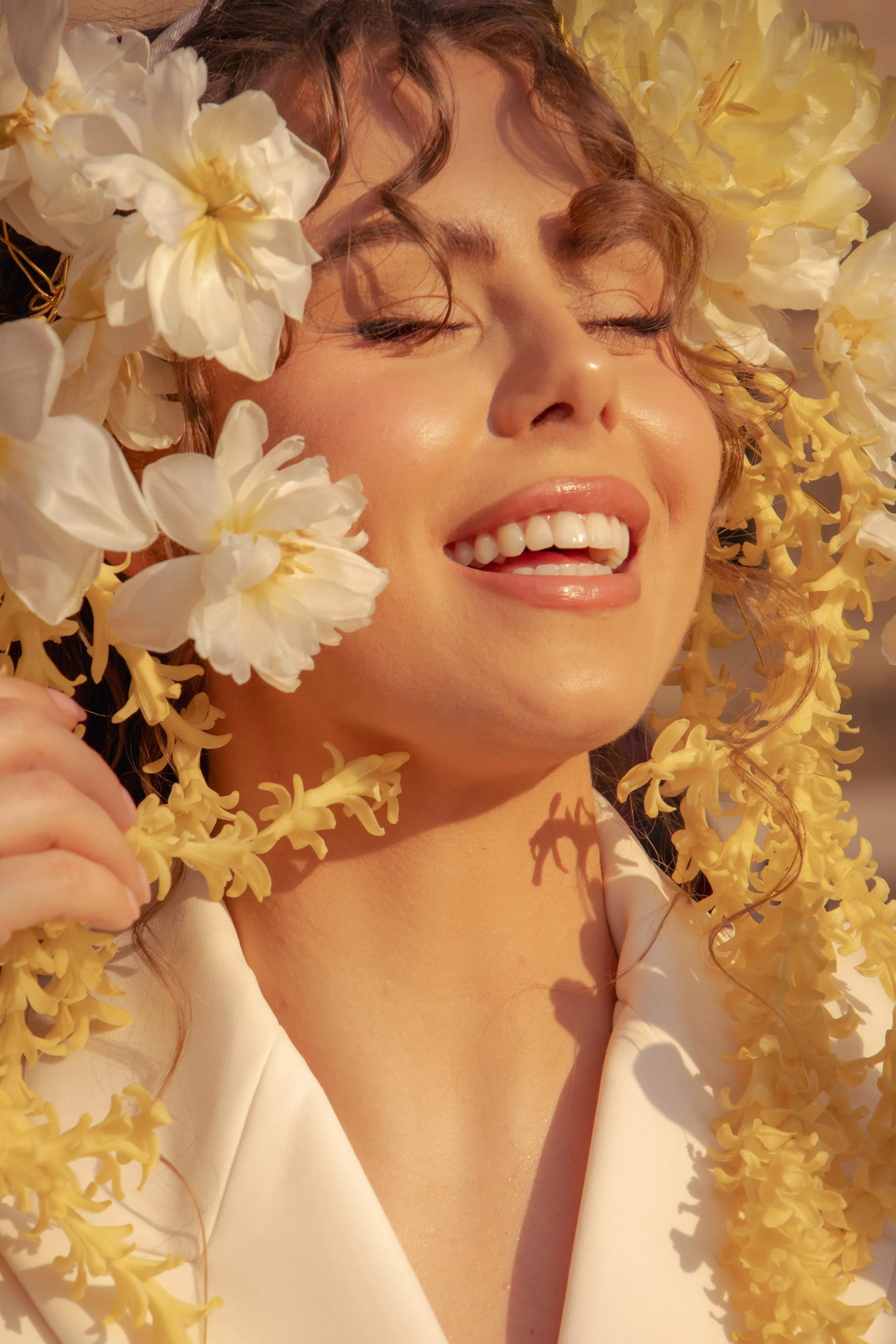A woman with curly hair and closed eyes smiling, surrounded by yellow and white flowers, wearing a white outfit.