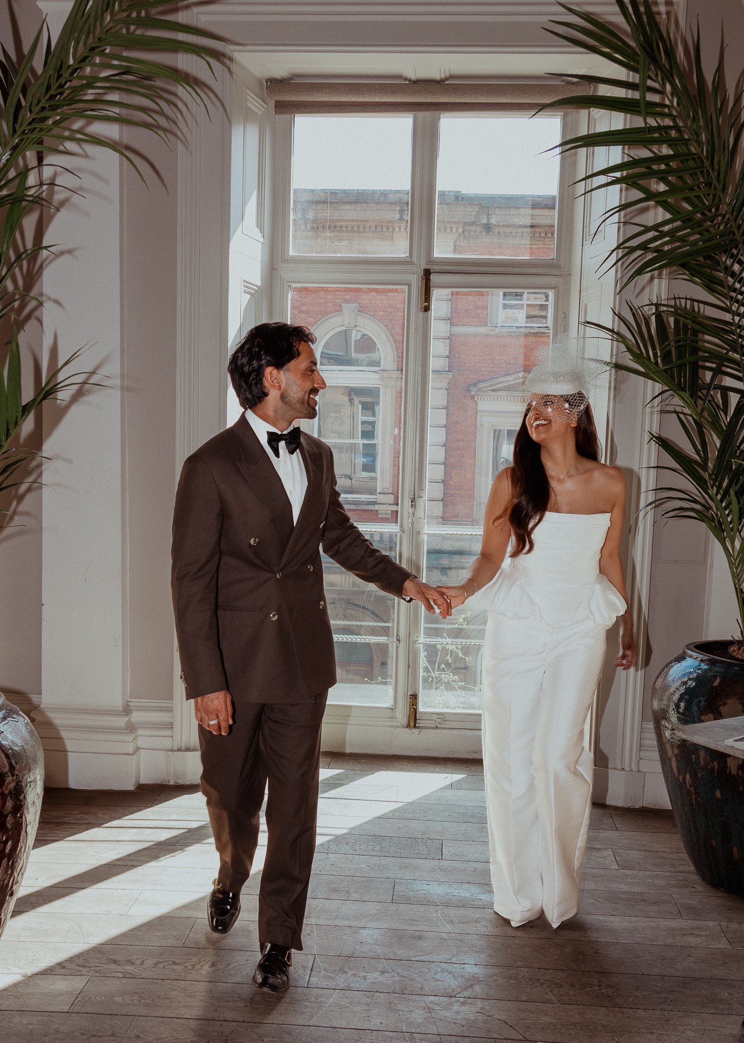 A man in a tuxedo and a woman in a white dress holding hands and smiling in front of large window with a cityscape view inside a well-lit room.