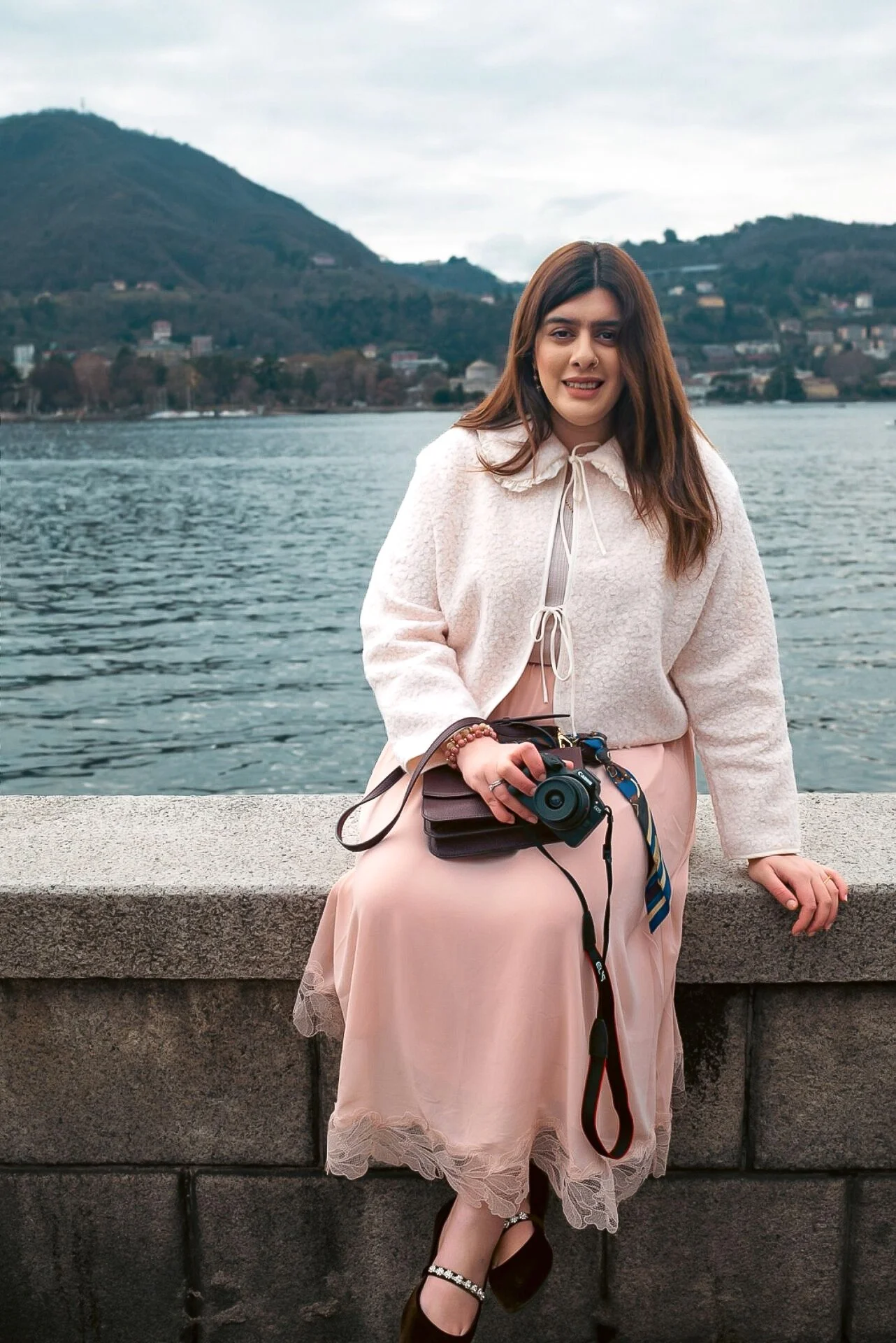 A woman with long brown hair sitting on a stone ledge by a body of water, holding a camera in her right hand, with a mountain and cloudy sky in the background.