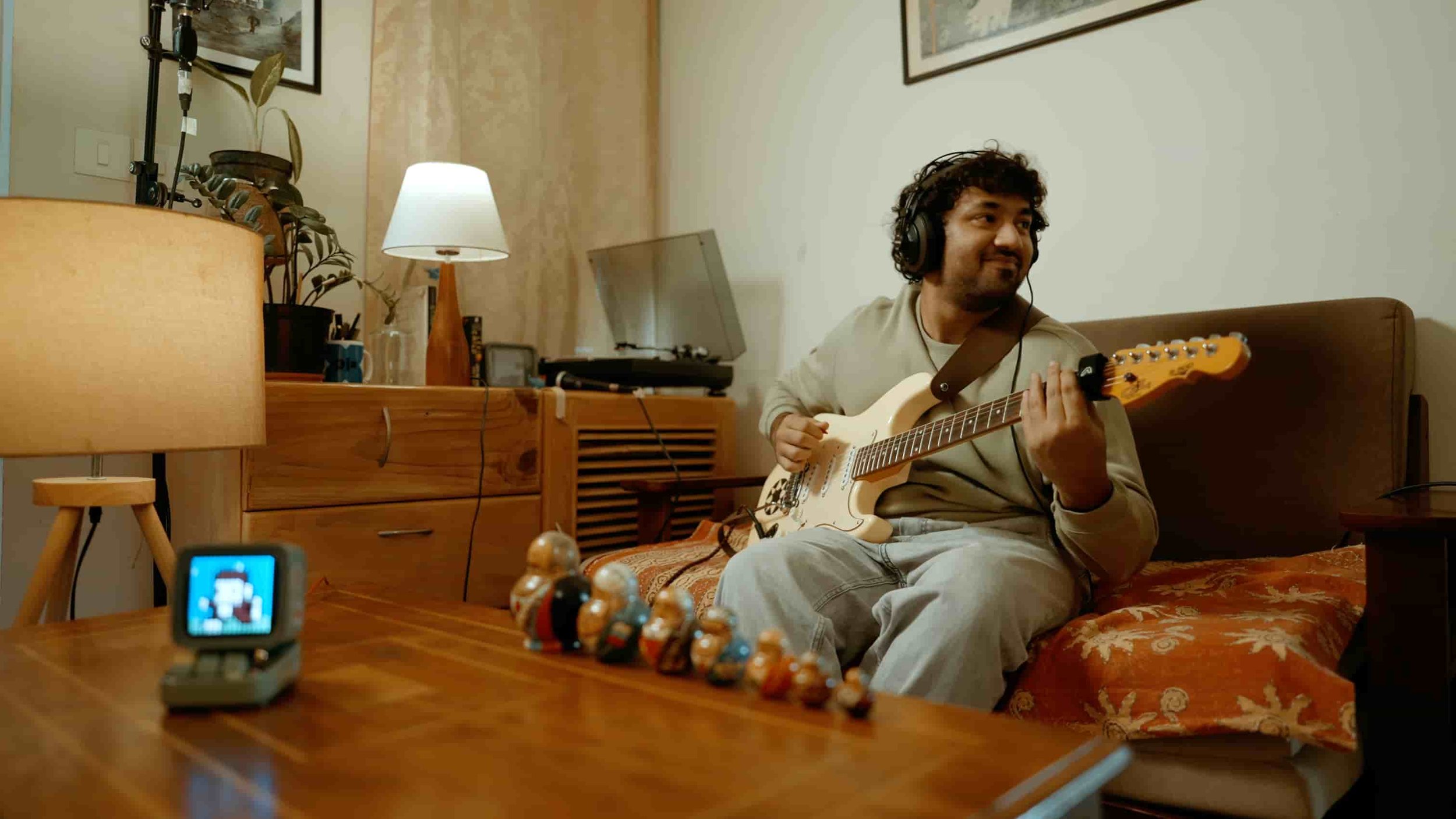 A man with curly hair and a beard, wearing a beige hoodie and gray sweatpants, sitting on a bed in a cozy room, playing an electric guitar. He is smiling and wearing headphones. The room has a wooden dresser with a lamp, a record player, and decorative plants, with warm lighting and framed pictures on the walls.