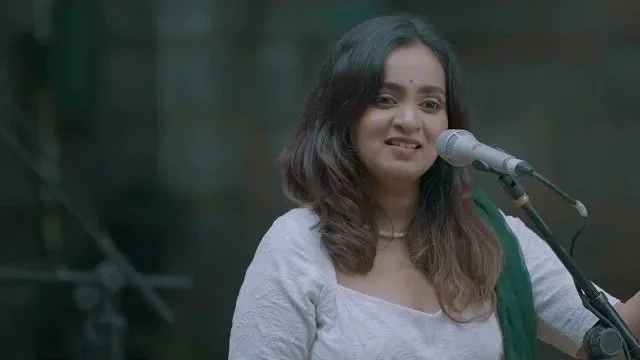 A woman with shoulder-length dark hair standing by a microphone, smiling slightly, wearing a white top and green dupatta, in an outdoor setting.