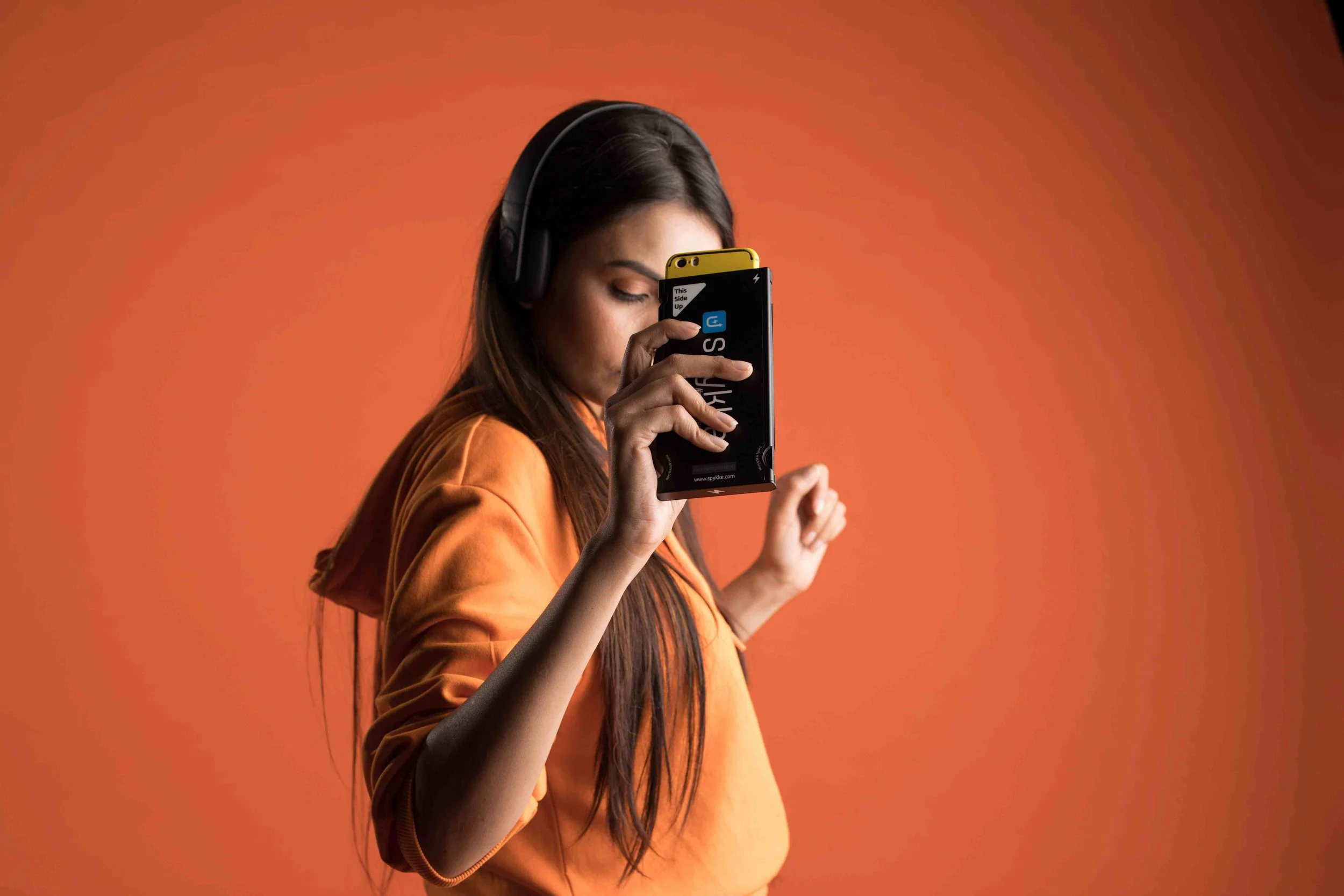 A young woman with long dark hair wearing an orange hoodie and black headphones, looking at her smartphone against an orange background.