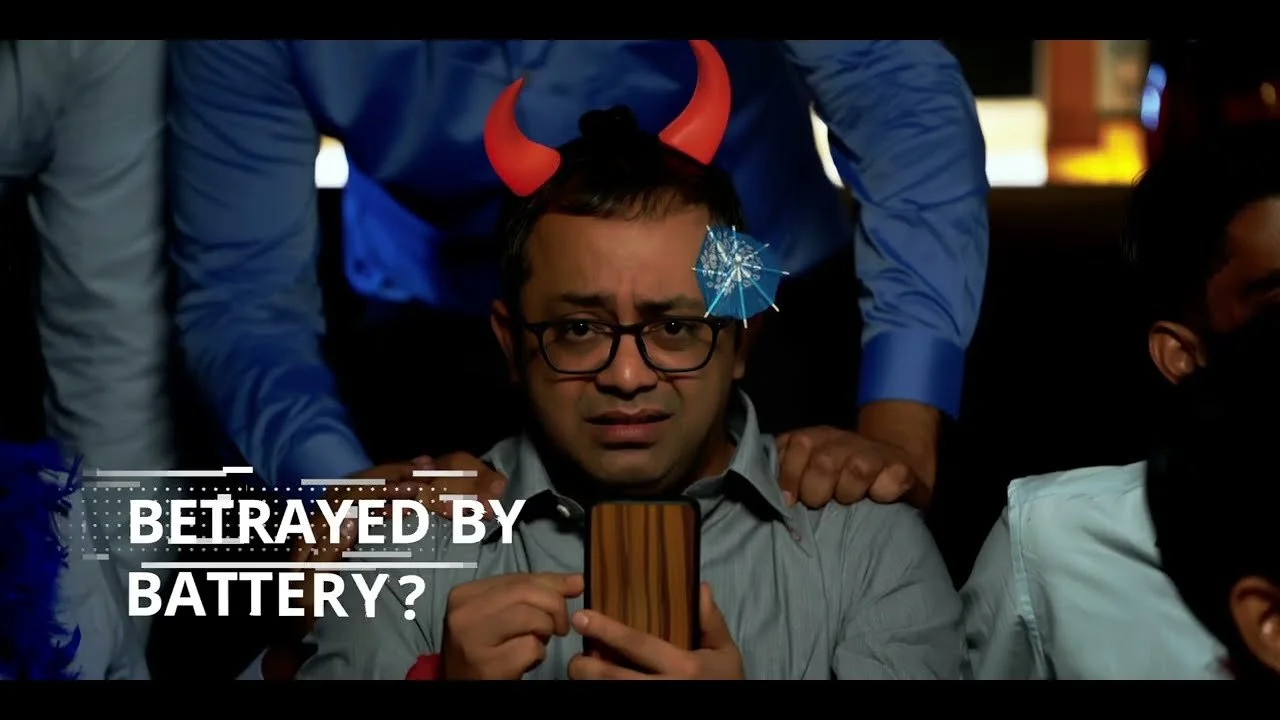 A man wearing glasses and a blue shirt is holding a smartphone, looking worried, with a person behind him wearing red devil horns and holding a small umbrella. The caption on the image reads "Betrayed by Battery?"