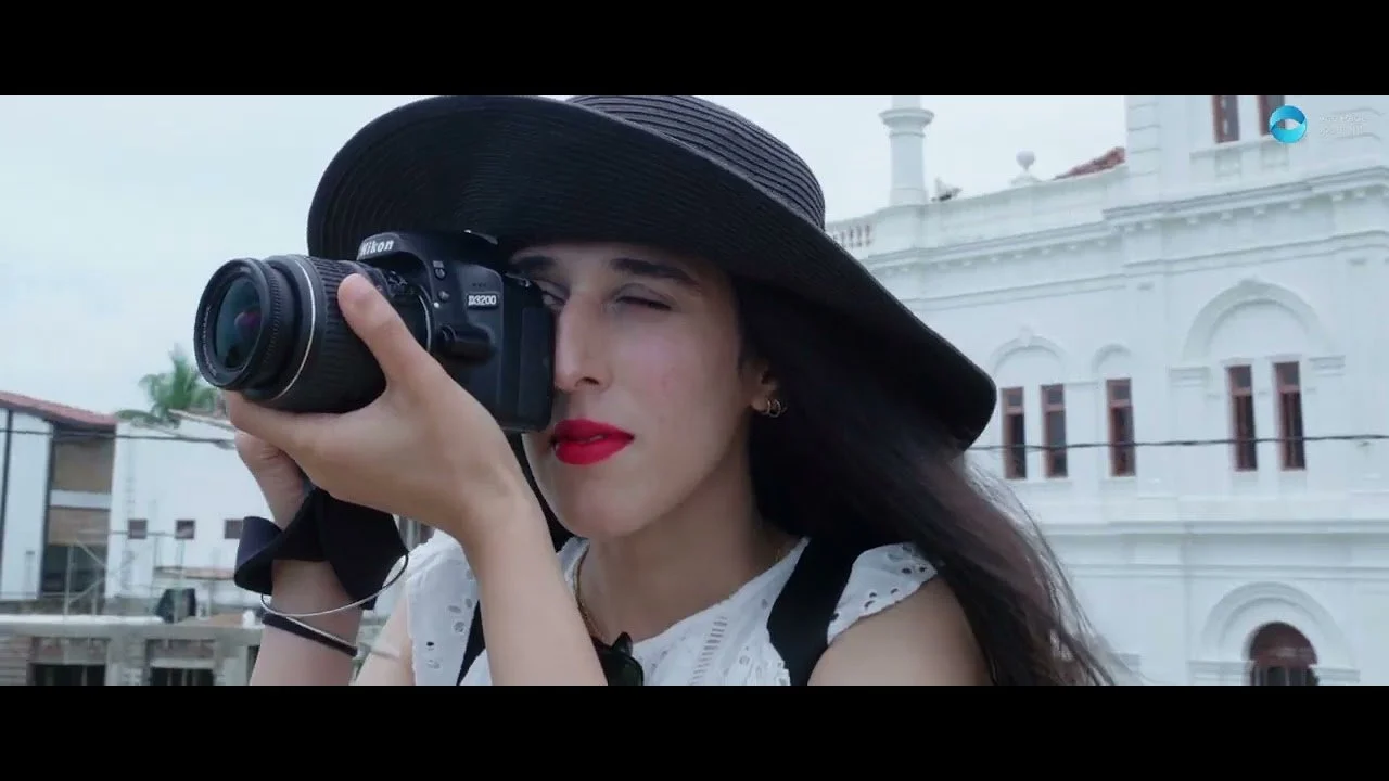 Woman with long dark hair, wearing a large black sun hat, takes a photograph with a DSLR camera, standing outdoors in front of a white building with architectural details.