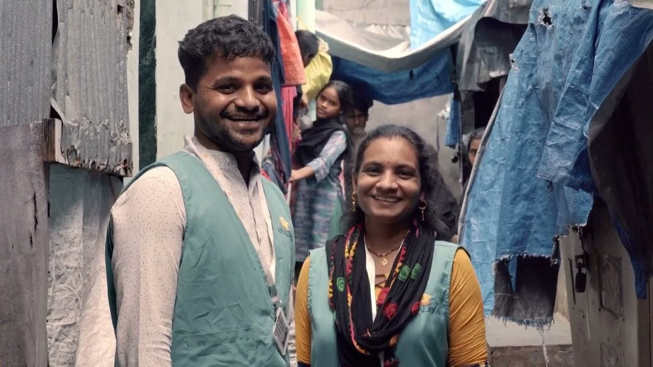 Smiling man and woman standing in a makeshift outdoor shelter with hanging clothes.