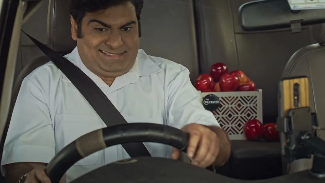 A man with a mischievous smile sitting in a vehicle with a seatbelt across his chest. There is a box of red apples behind him.