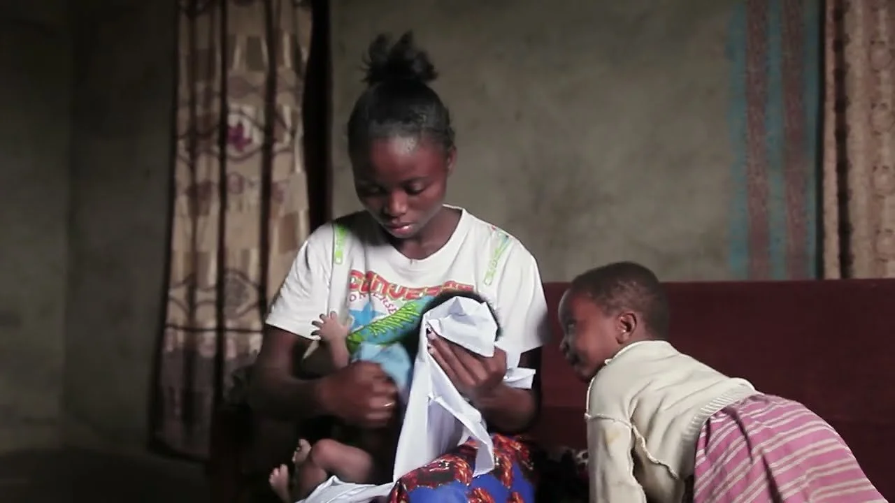 Young girl holding a baby, with a boy leaning on her on a sofa in a modest room.