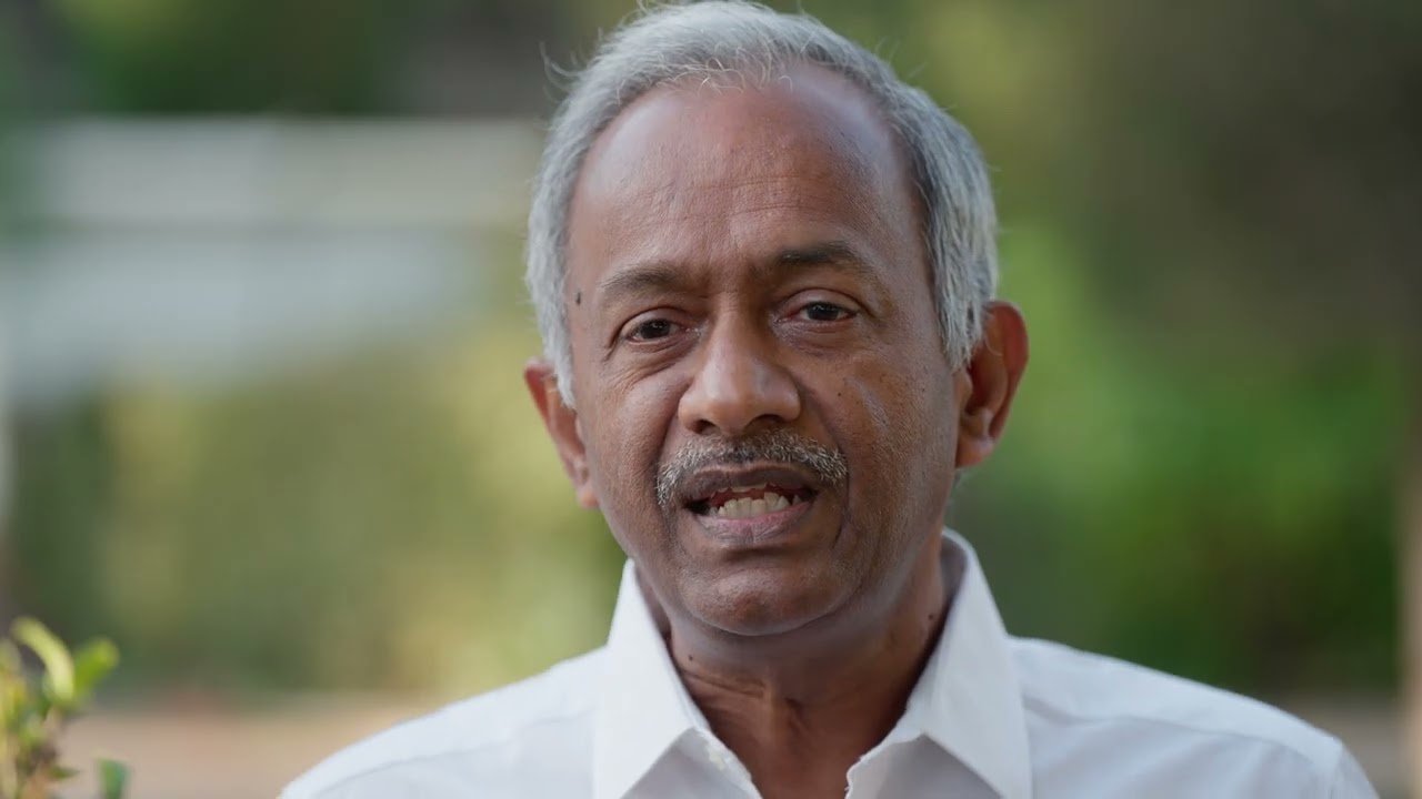 Close-up of an elderly man with gray hair and a mustache, speaking outdoors with a blurred green background.
