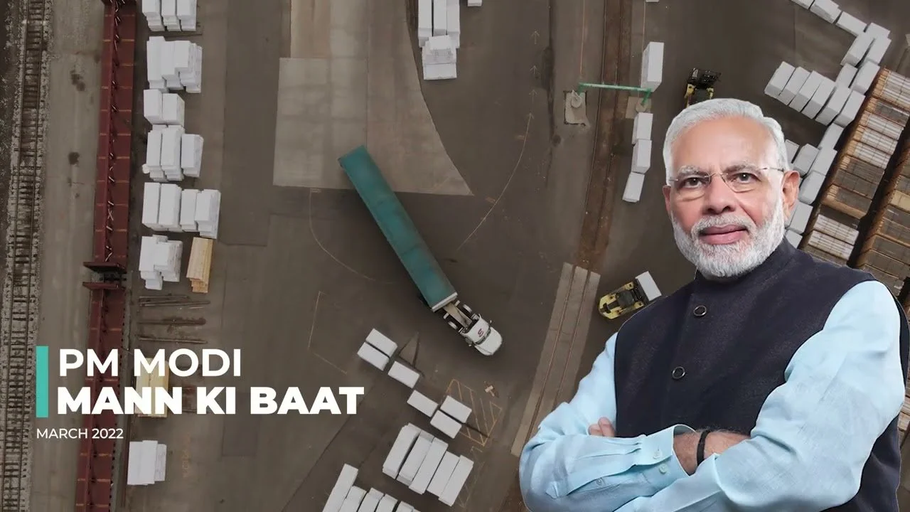 An aerial view of a construction site with trucks, pallets, and building materials. In the foreground, a man with white hair and beard wearing glasses, a light blue shirt, and a dark vest is smiling and crossing his arms.