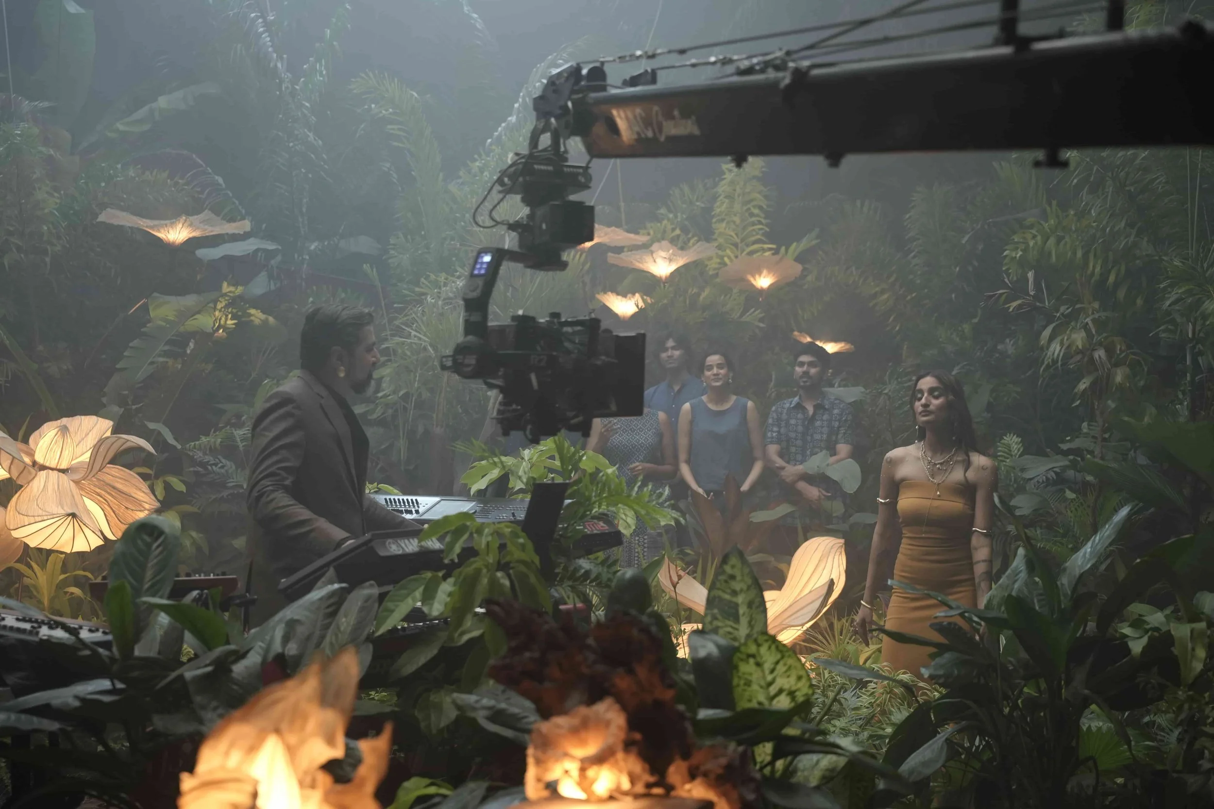 Filming scene inside a lush, misty jungle with foliage and illuminated flowers, showing a director and a crew filming a woman in a tan dress among plants.