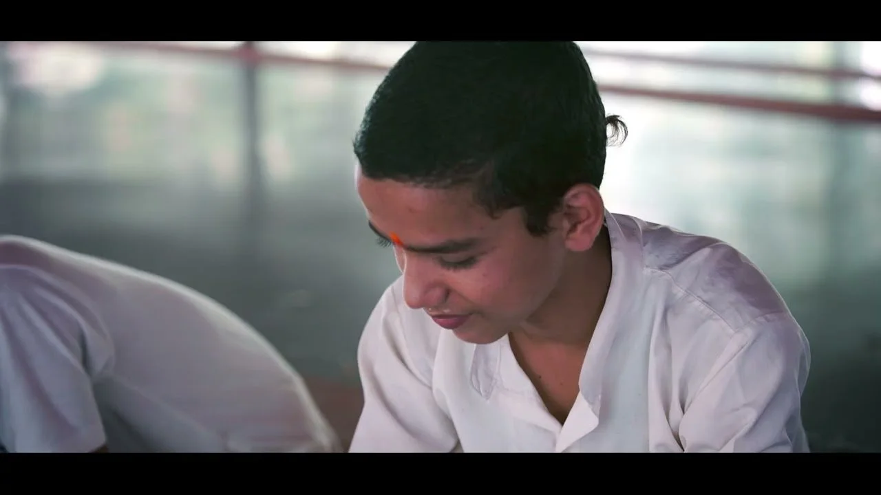 A young man in a white shirt looks downward, with a slight smile in a softly lit indoor setting.