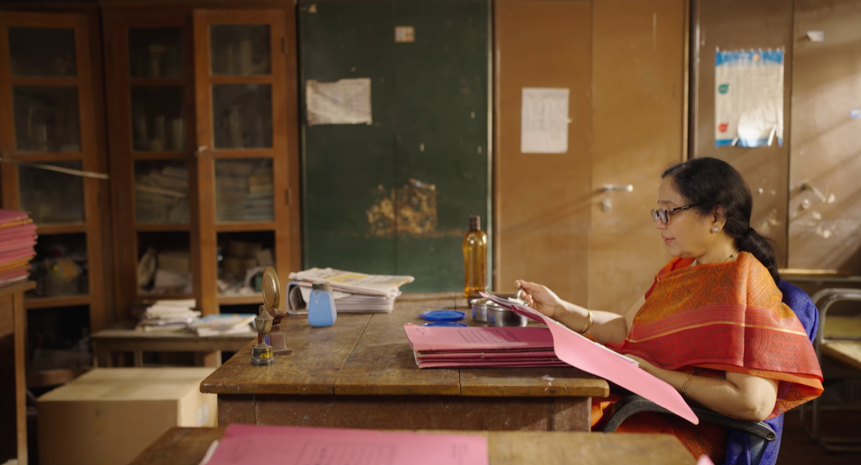 A woman wearing glasses and a red saree sitting at a cluttered wooden desk with pink file folders and papers, looking at a document in her hand in an office with wooden cabinets and a blackboard.