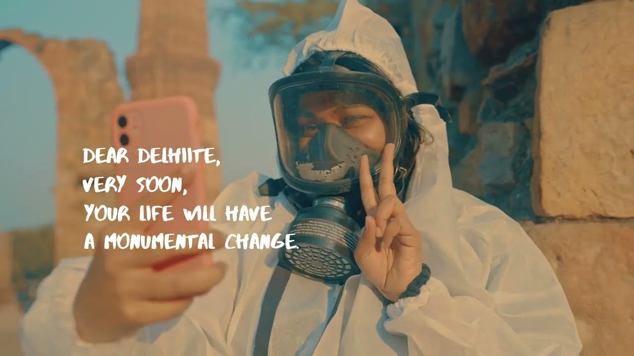 A person wearing protective gear, including a hazmat suit, mask, and goggles, taking a selfie with a pink phone against a backdrop of ancient stone ruins.