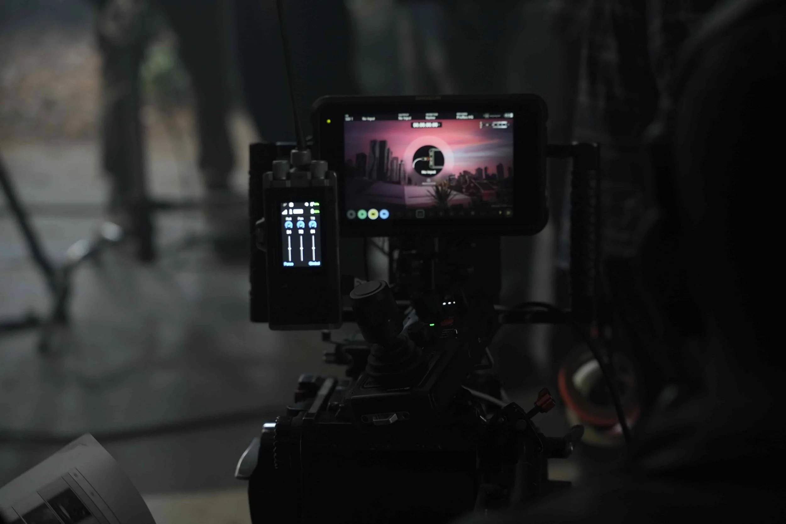 Close-up of a professional video camera with an external monitor displaying a sunset cityscape scene, set up in a dim environment.