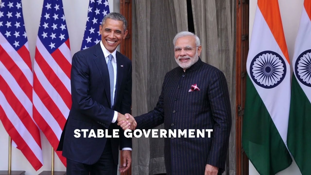U.S. President Barack Obama and Indian Prime Minister Narendra Modi shaking hands and smiling, with flags of the United States and India in the background, in a diplomatic meeting.