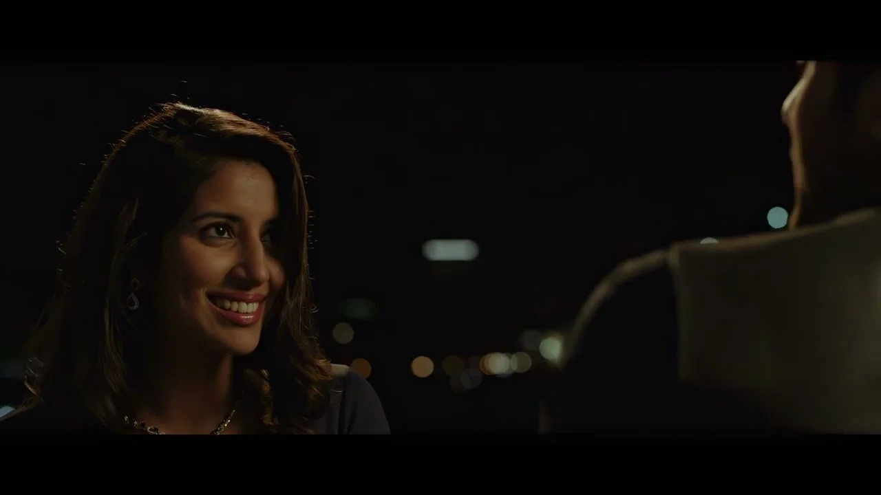 A woman smiling at a man during a nighttime conversation, with city lights blurred in the background.