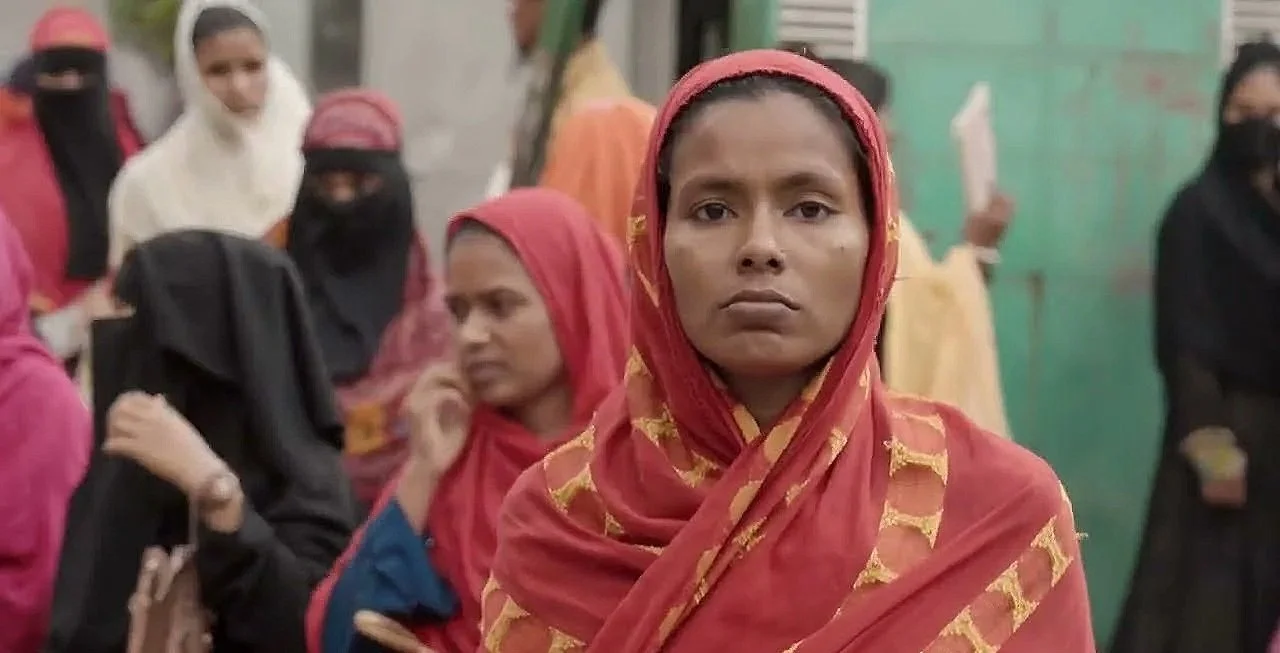 A woman in the foreground with a serious expression, wearing a red and gold headscarf and traditional clothing. Behind her, several women are wearing headscarves and face coverings, with some reading or looking at something. The background shows a green wall.