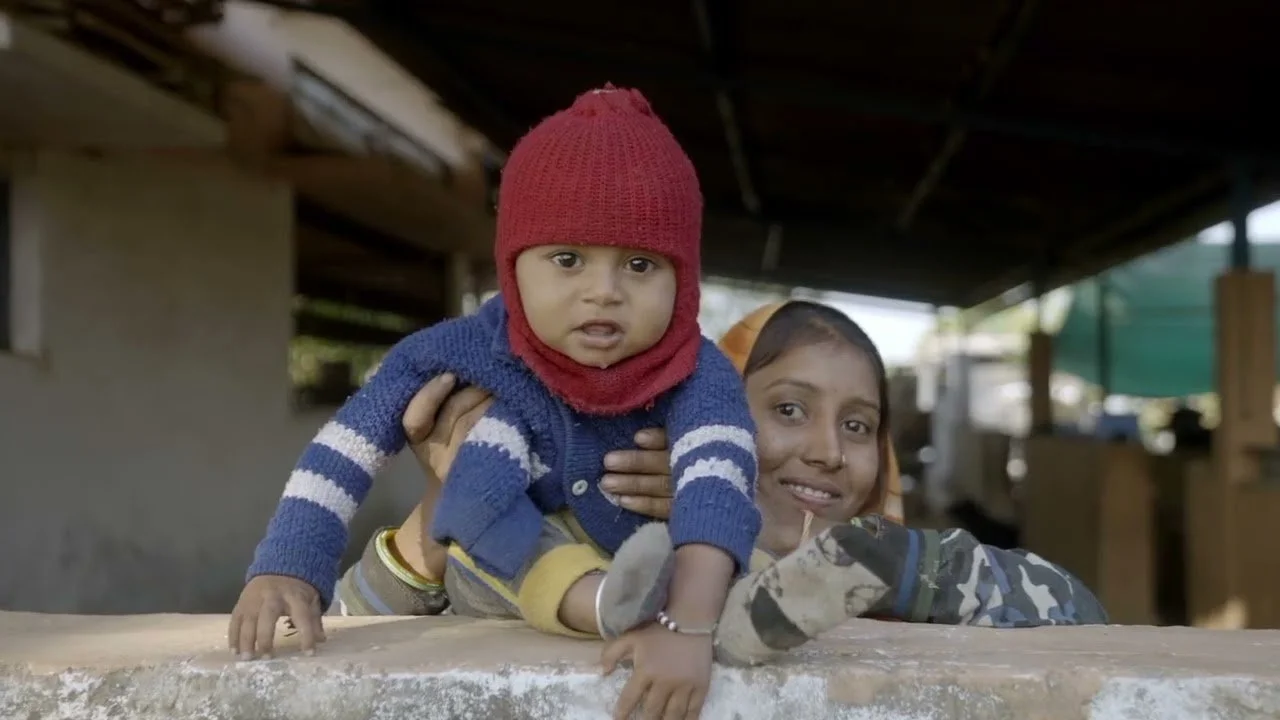 A woman holding a young child with a red knit cap in an outdoor setting, smiling at the camera.