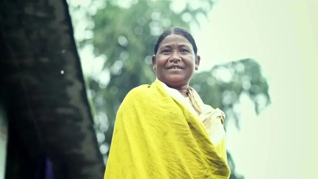 Smiling woman wearing a yellow saree outdoors with blurred trees in the background.