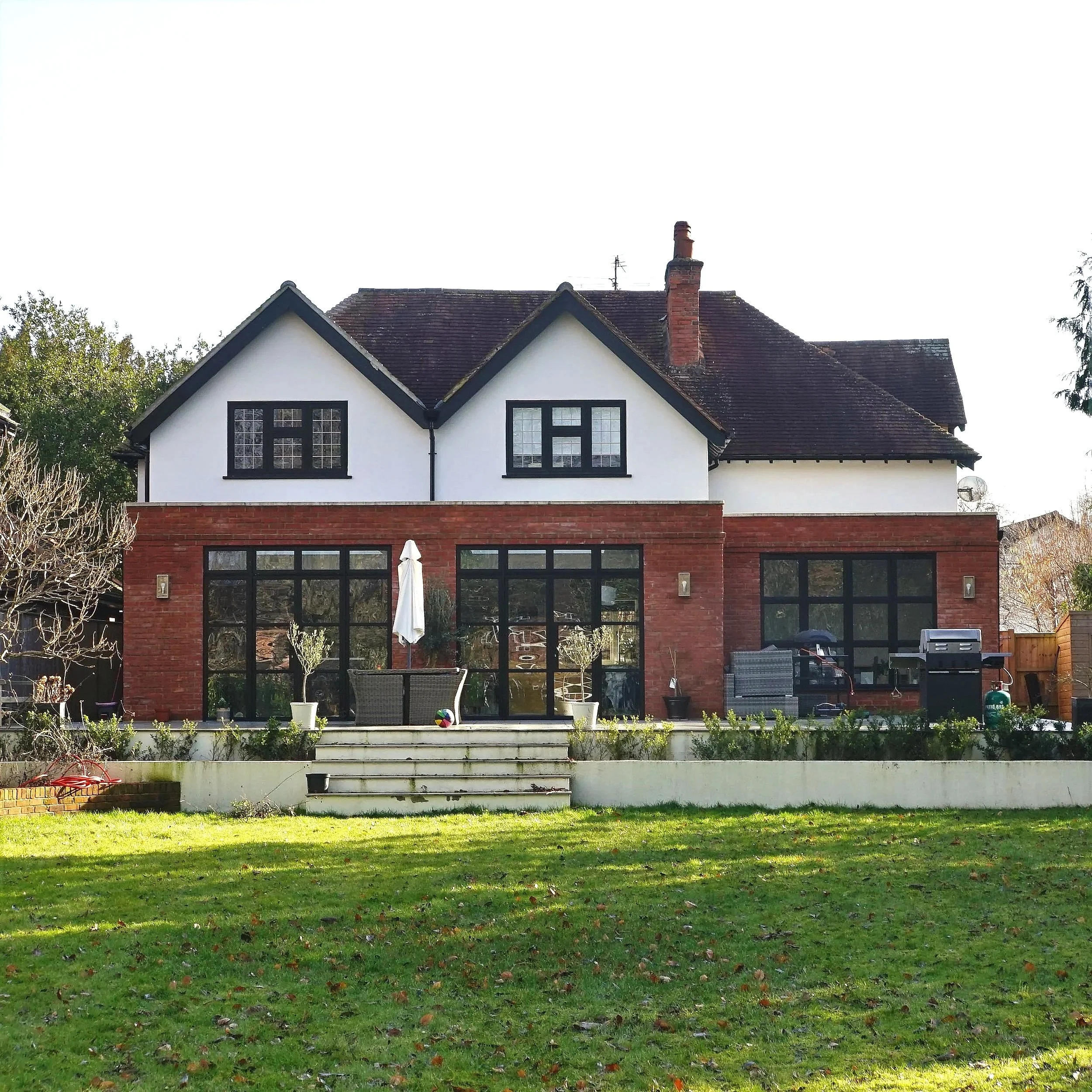 Large modern house with white walls, black window frames, and a sloped roof with red tiles. The backyard features a patio area, green lawn, and garden beds.