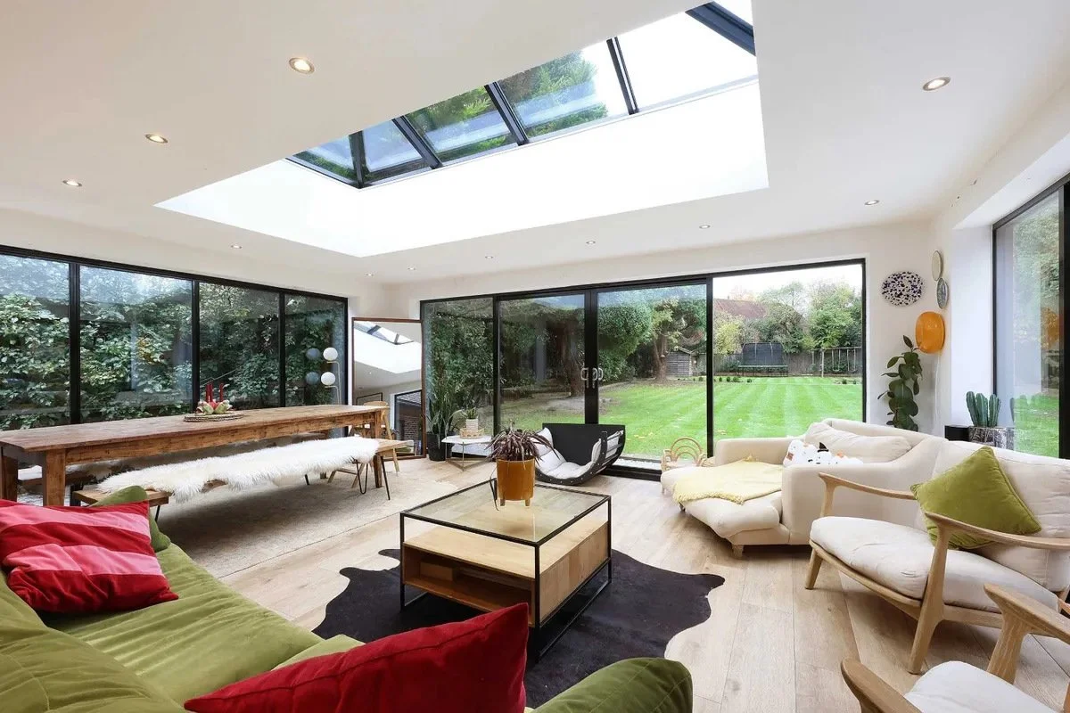 Modern living room with large glass sliding doors opening to a green backyard, featuring a skylight, white and beige sofas, a wooden coffee table, and decorative wall art.