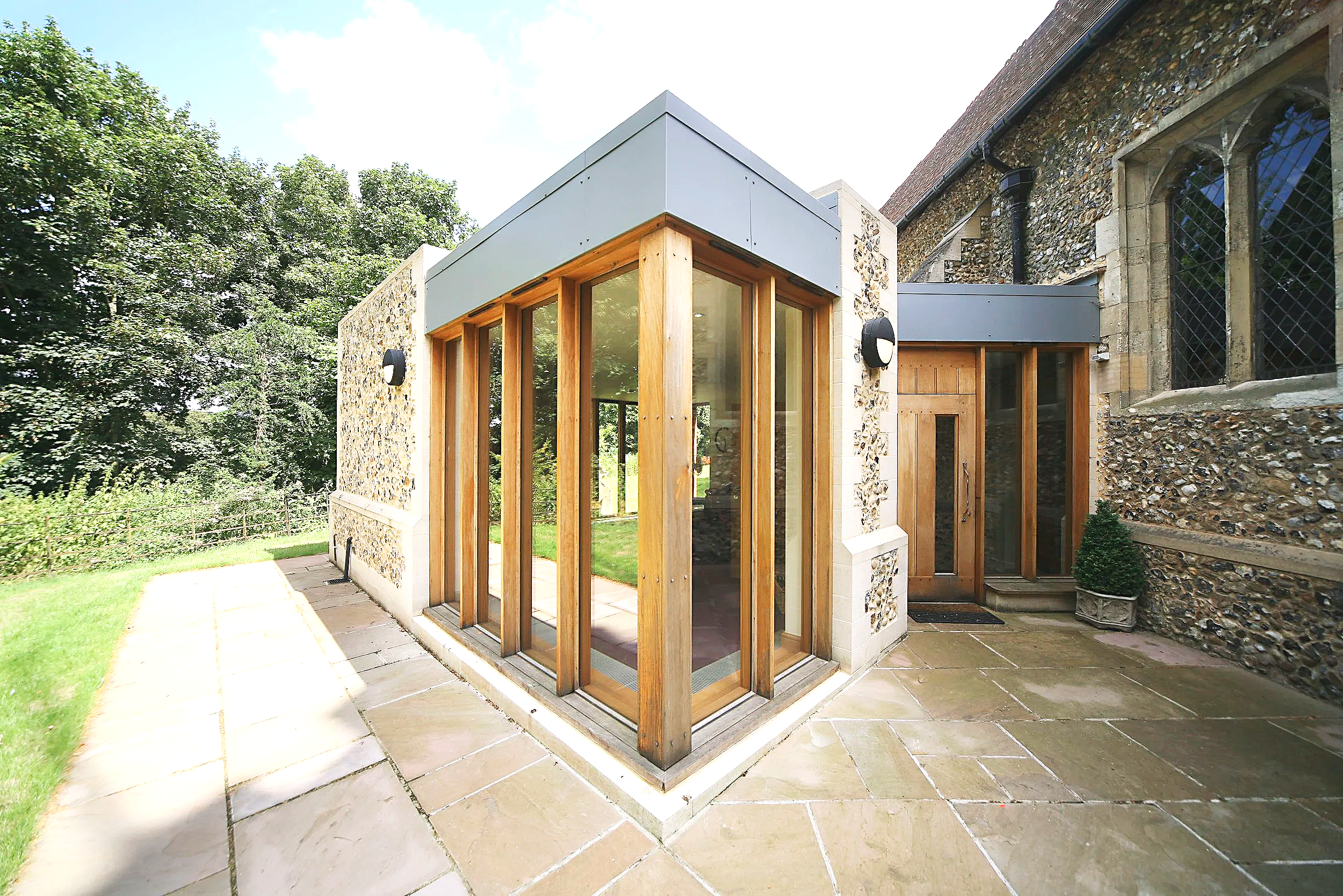 Exterior view of a modern church extension with large glass windows, oak window framing, and flintwork walls, attached to a historic church building with flintwork walls and stained glass windows, surrounded by greenery and paved patio.