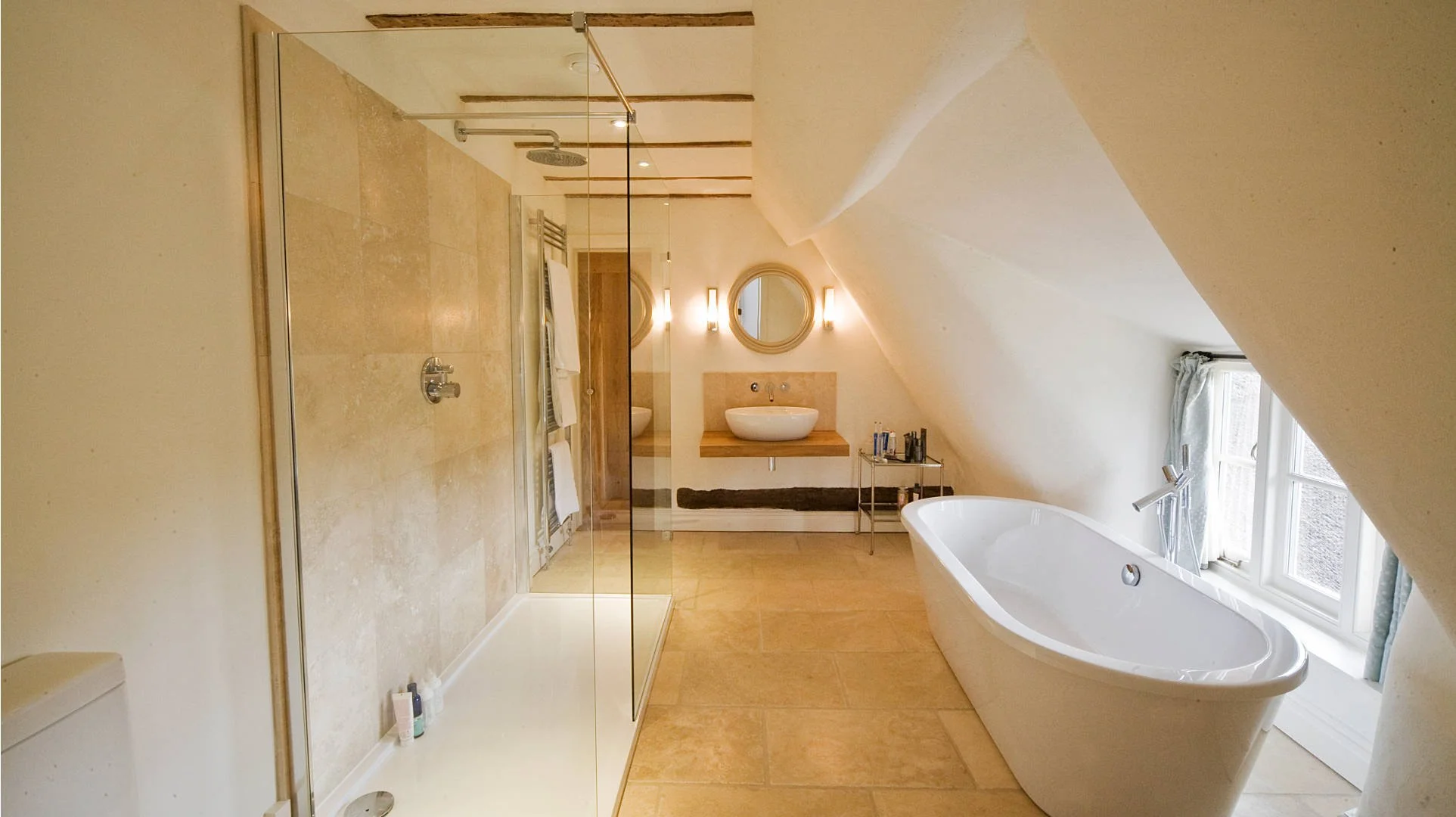 A bathroom with a glass-enclosed shower, a freestanding bathtub by a window, and a wall-mounted sink with a round mirror above. The room has beige tiles and walls, a small metal cart with toiletries, and wall-mounted lights.
