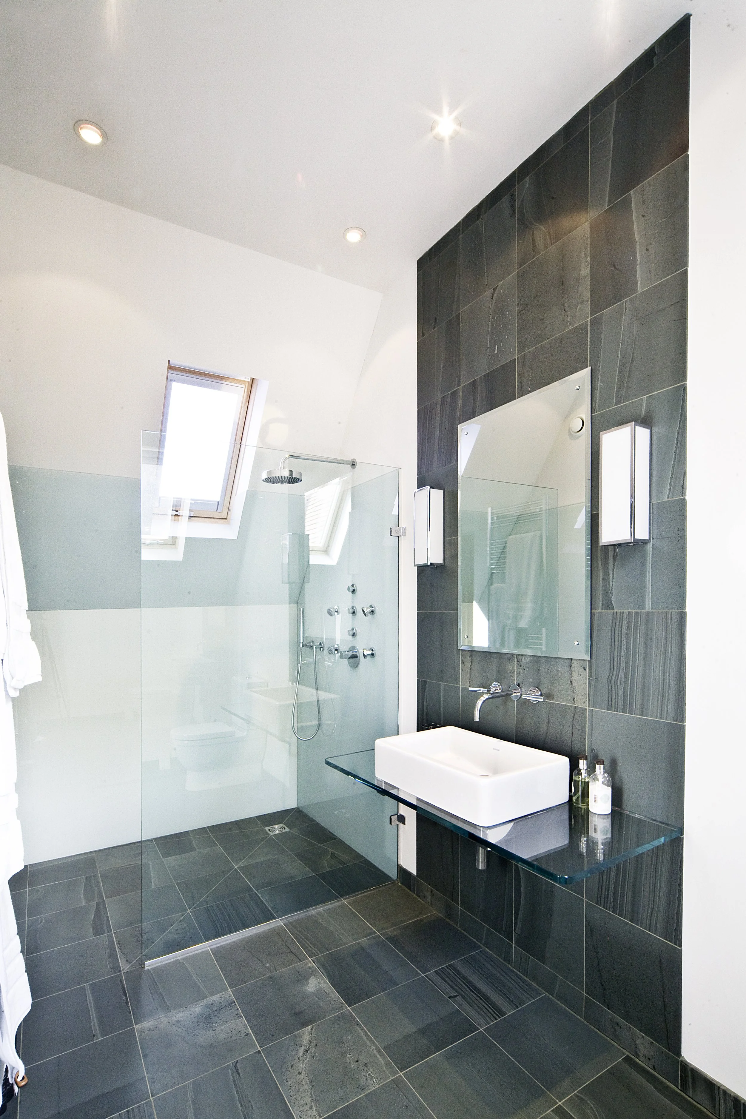 Modern bathroom with dark gray tiled wall and floor, glass shower enclosure with a skylight in the ceiling, mirror, white sink, and various toiletries.