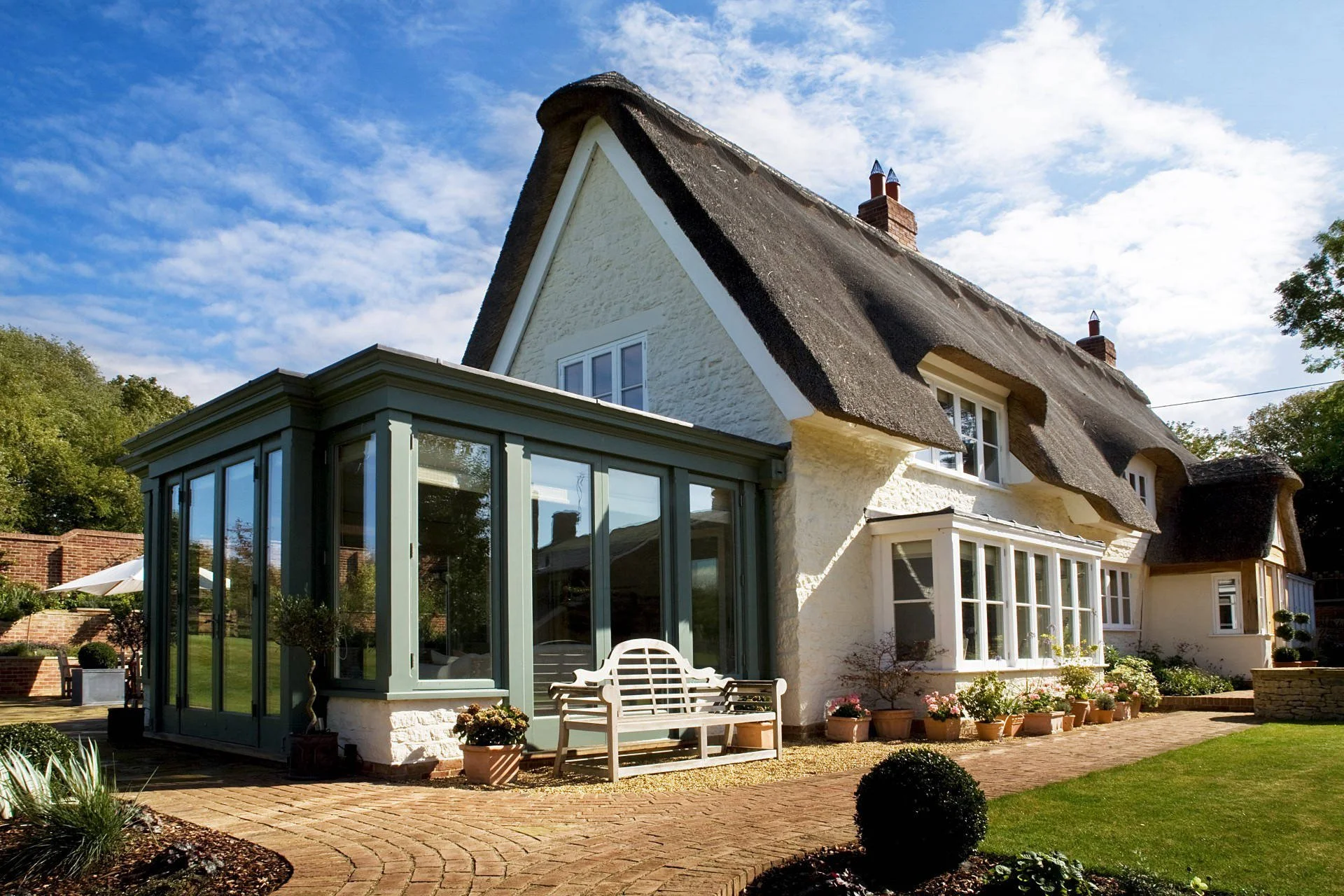 A house with a thatched roof and white exterior walls features a glass-enclosed sunroom with a pale green frame, a white wooden bench, and potted plants in the garden. The sky is blue with scattered clouds, and there is a brick patio with garden beds and a lawn.