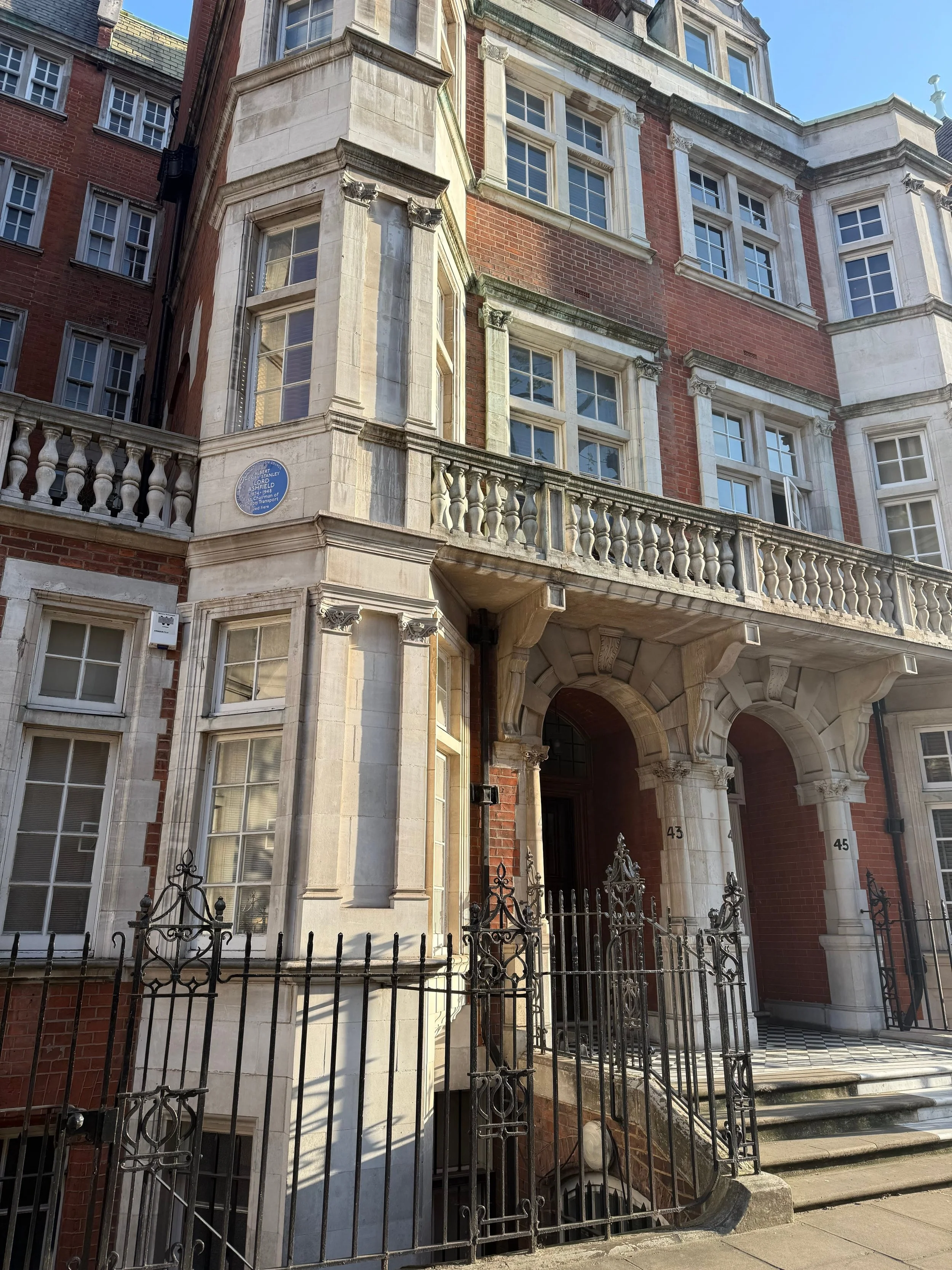 A historic brick building in Mayfair, London with ornate architectural details, including large windows with white frames, a stone balcony railing, and decorative stonework around the entrance. A black wrought iron fence surrounds the front steps.