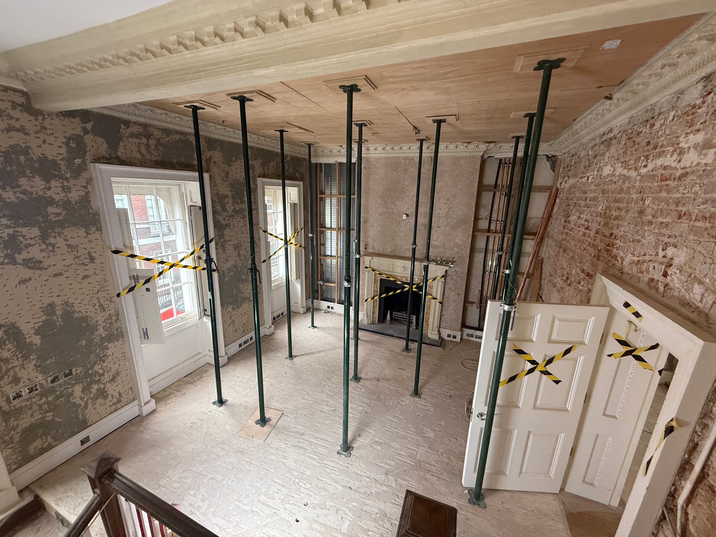 Room under renovation with scaffolding poles, unfinished walls with patches of exposed brick and plaster, wooden flooring, and large windows with white molding.