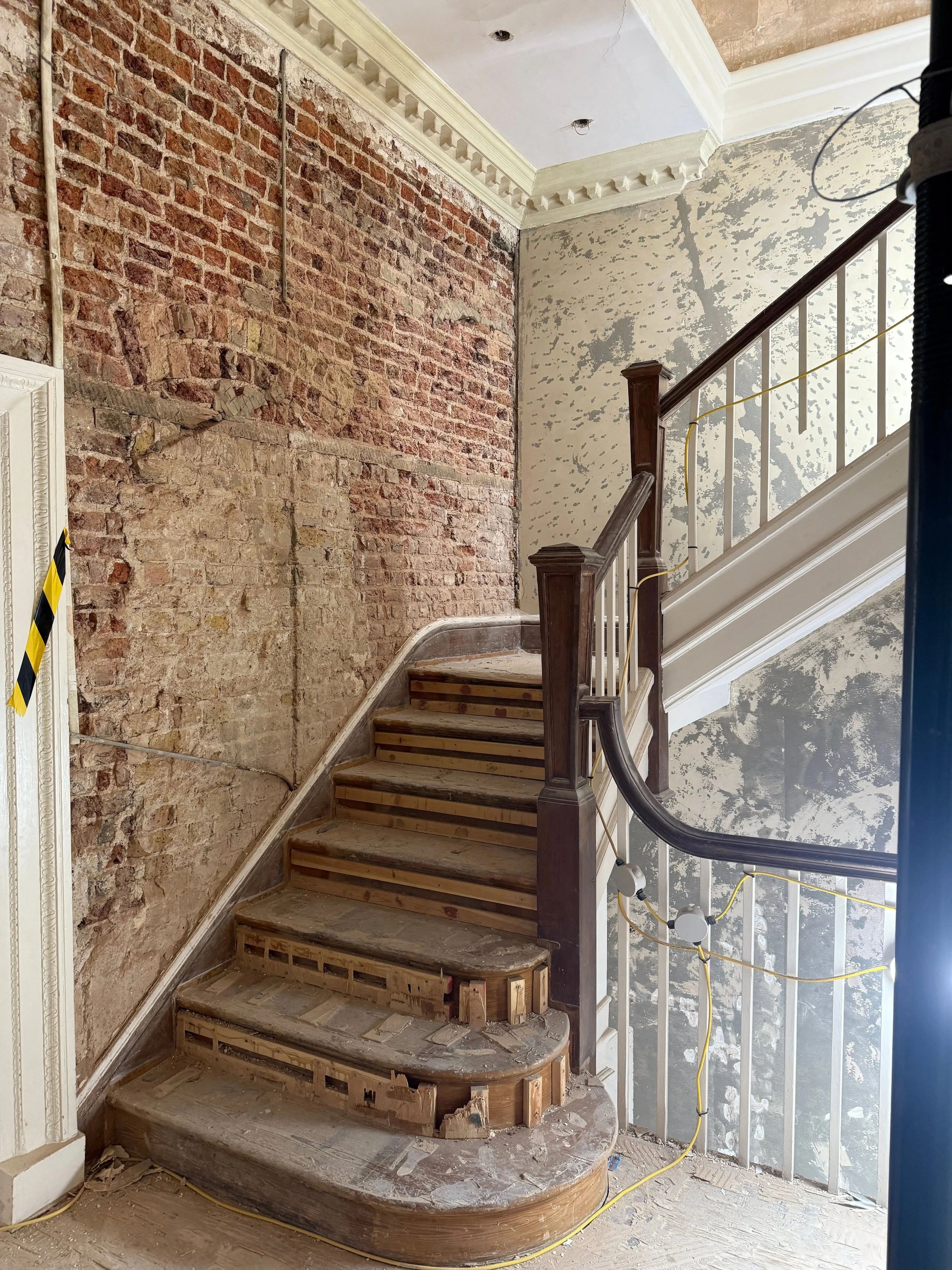 A staircase under renovation with exposed brick and drywall, construction materials, and electrical wiring visible.