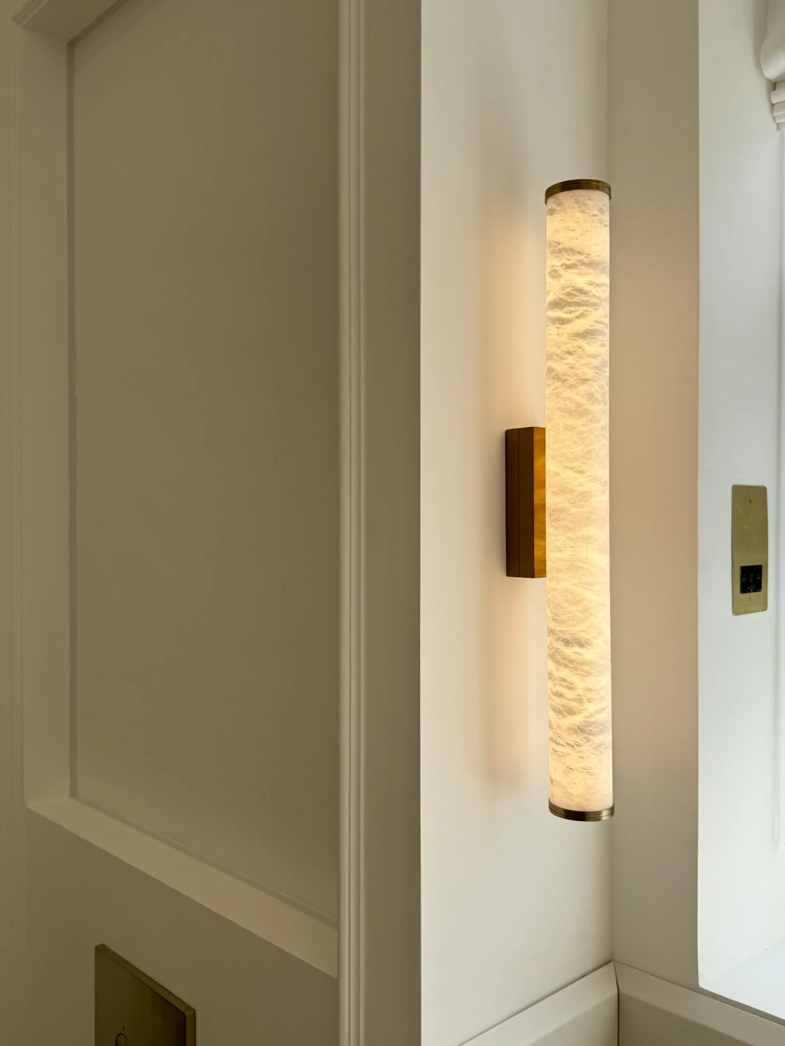 Close-up of a modern wall-mounted cylindrical light fixture with a textured surface, attached to a white wall near an electrical outlet.
