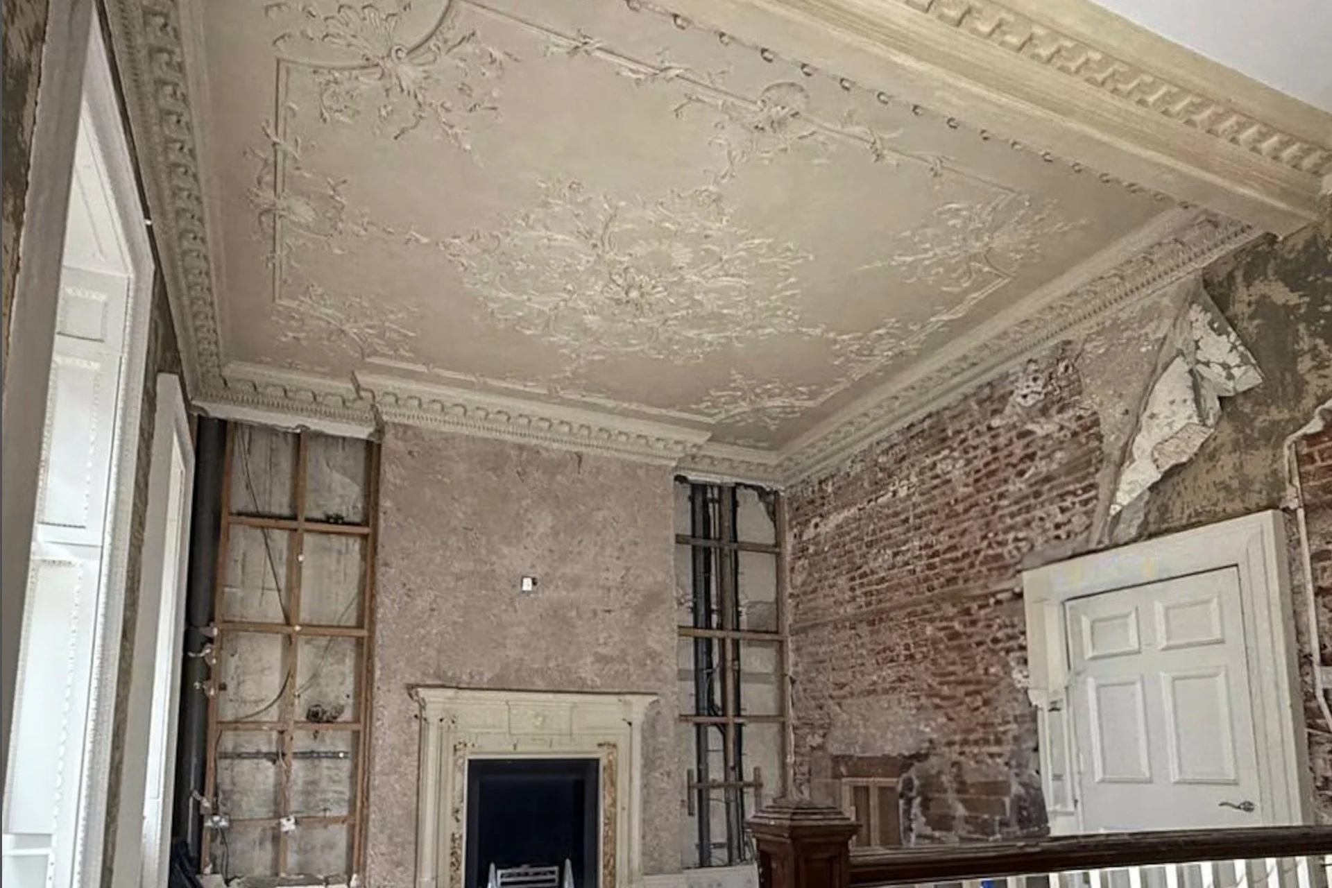 Interior of a room under renovation with exposed brick walls, partially removed plaster, decorative ceiling molding, and a white door.