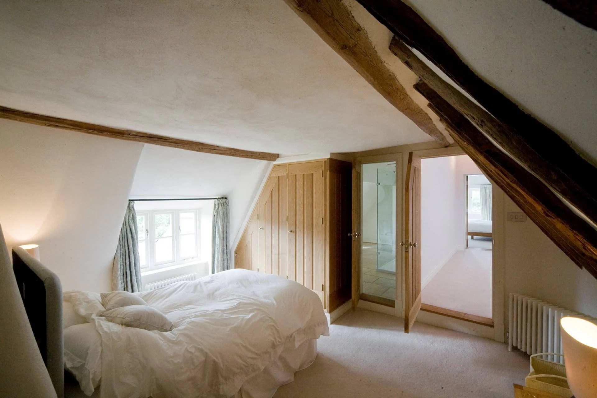 A cozy bedroom with a sloped ceiling and exposed wooden beams. The bed has white bedding and is positioned near a window with curtains. There is a wooden wardrobe and a glass-enclosed shower visible in an adjacent room.