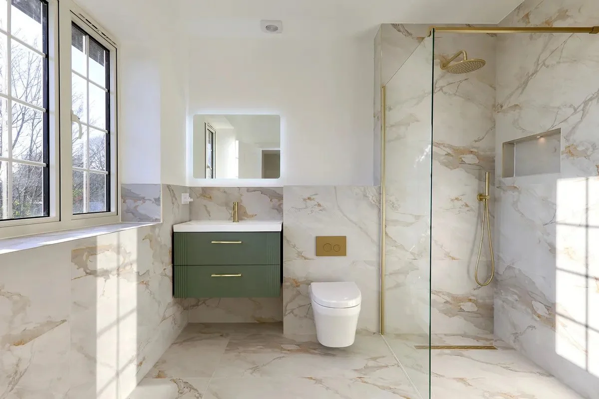 Modern bathroom with marble tile walls and floors. Features a large window, green vanity with gold handles, wall-mounted toilet, and a walk-in shower with a gold showerhead and glass partition.