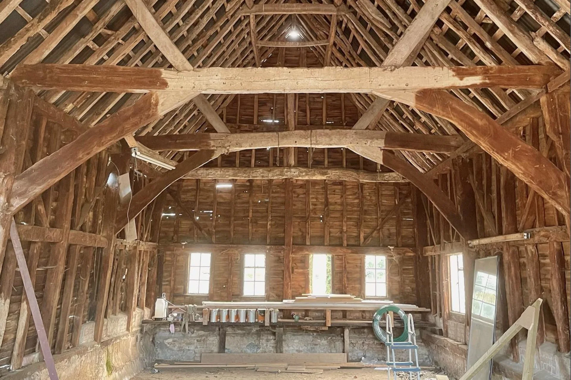 The interior of an old barn under renovation, showing exposed wooden beams, rafters, and windows.