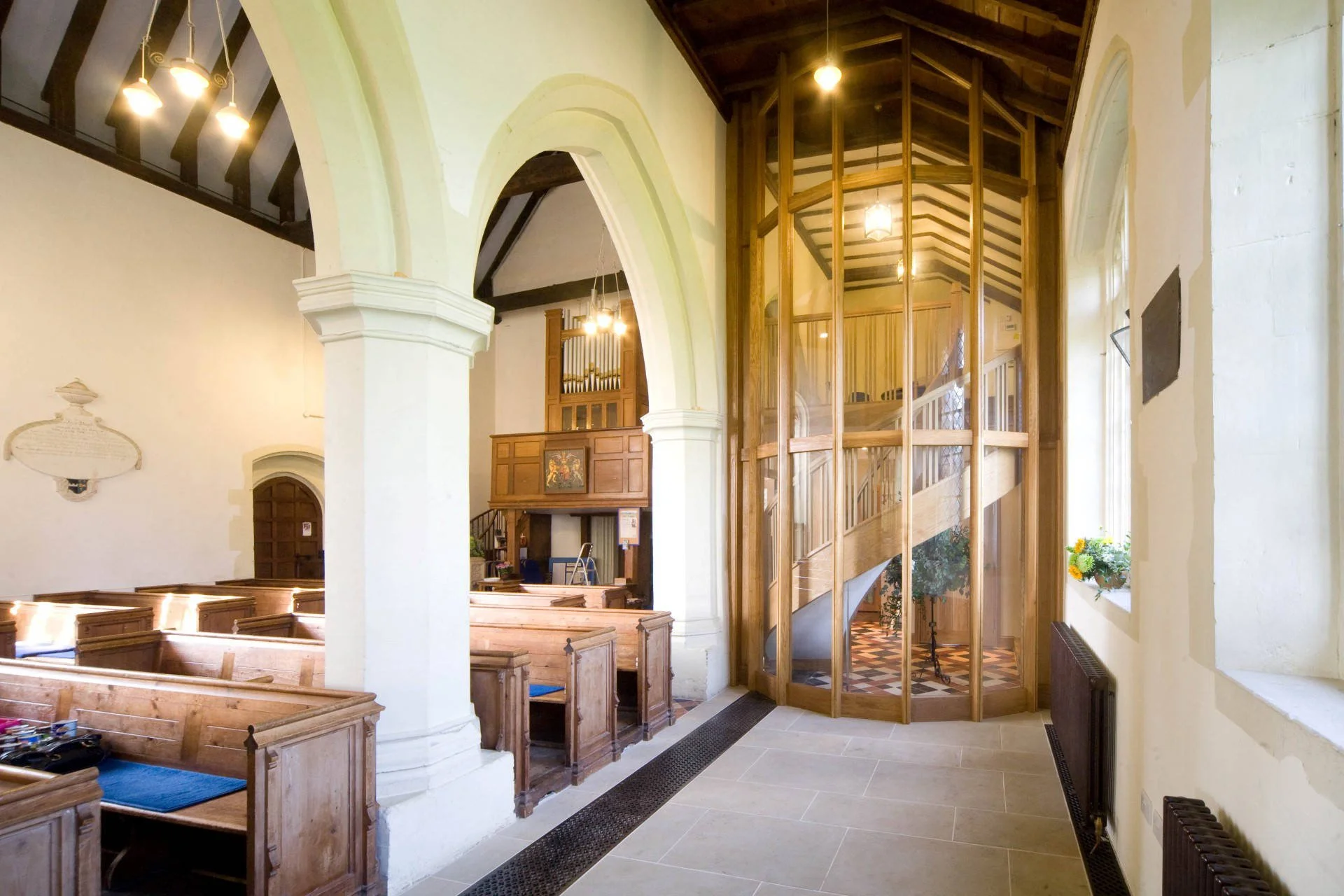 Interior of a church with wooden pews, ornate architecture, and a wooden and glass balcony, adorned with chandeliers and floral arrangements.