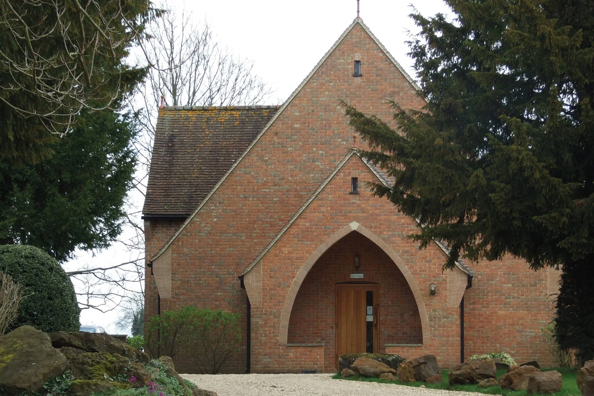 A brick church hall with an arched entrance, surrounded by trees and shrubs, with a wooden door and small windows. The roof is sloped and covered with clay plain tiles.