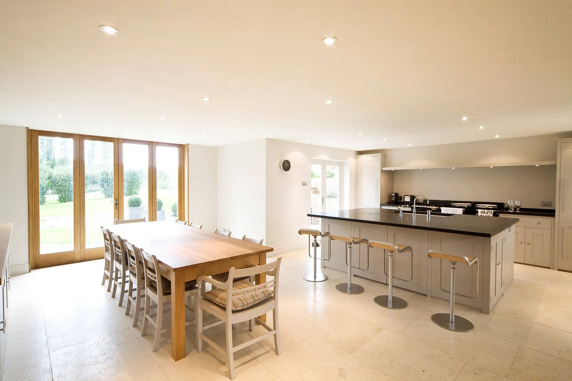 Bright, open kitchen and dining area with large wooden table, white chairs, and a kitchen island with bar stools. Features large windows and doors leading outside, recessed ceiling lights, and white cabinetry.