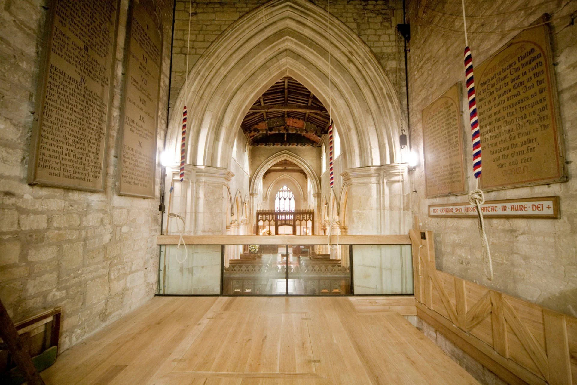 Interior view of a church with stone walls and high arched gothic windows. Wooden flooring, large religious plaques with text on the walls, and American flags hanging vertically on either side of the arch.