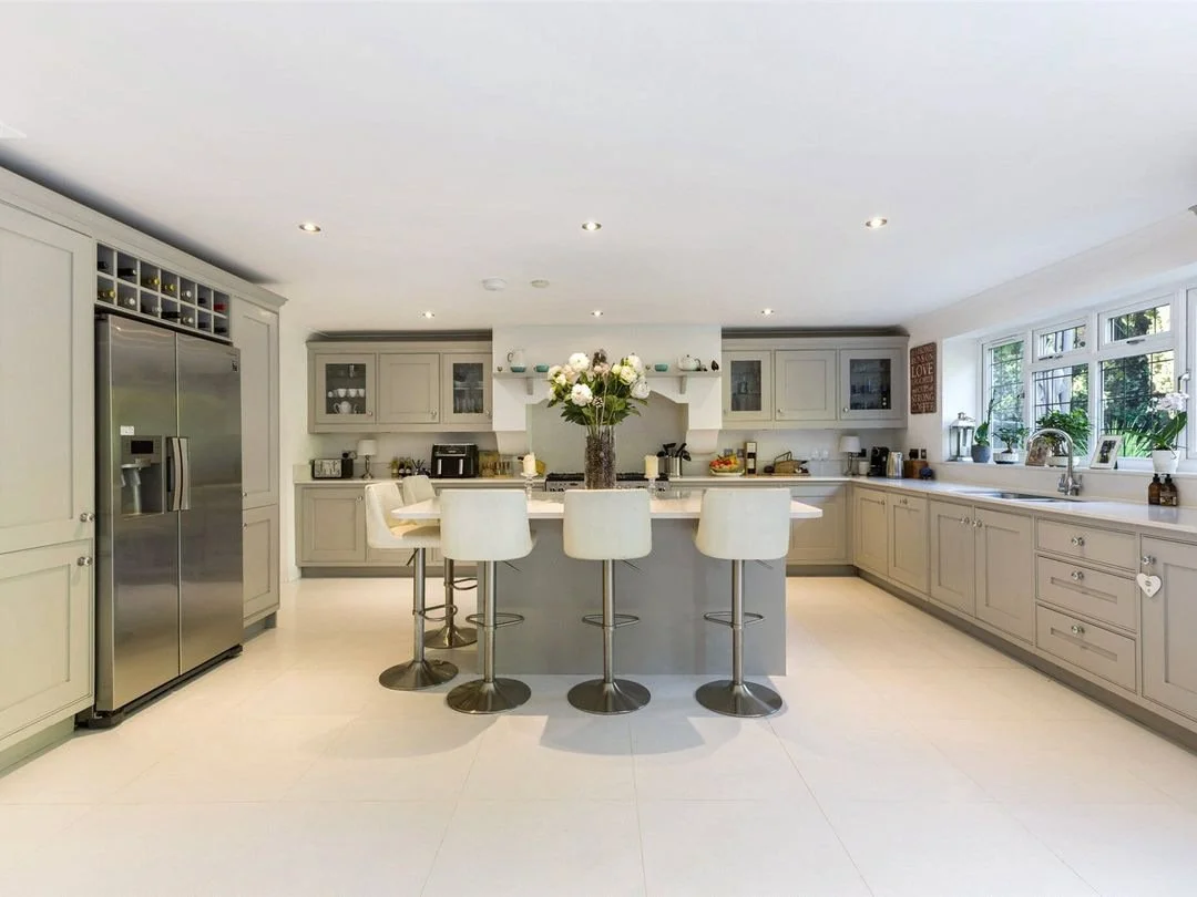 Bright, modern kitchen with light gray cabinetry, stainless steel appliances, a large island with white bar stools, and a window overlooking outdoor greenery. Central vase with white flowers on the island.