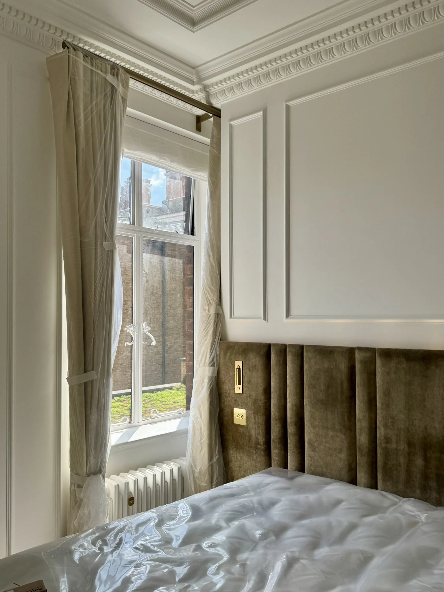 Interior of a room with a window, cream curtains, and a padded headboard, showing a bed with white bedspread and a view of neighboring buildings and blue sky outside.