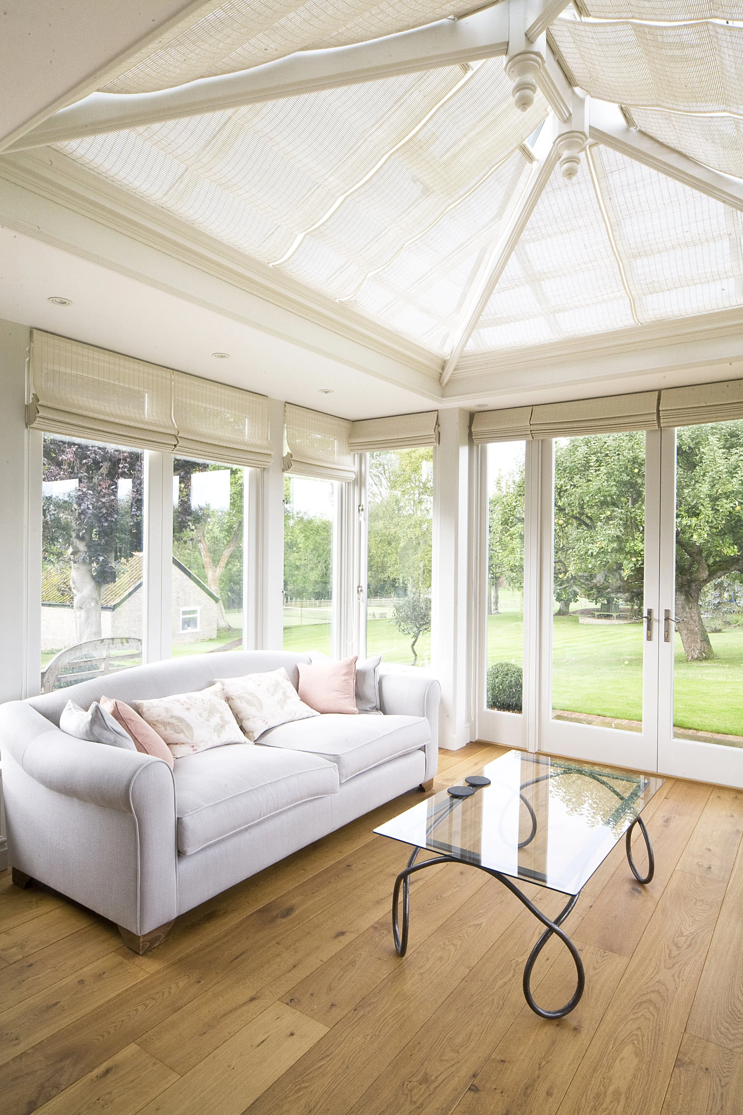 Bright sunroom with large windows, a white sofa with pillows, a glass coffee table, and a view of a green lawn outside.