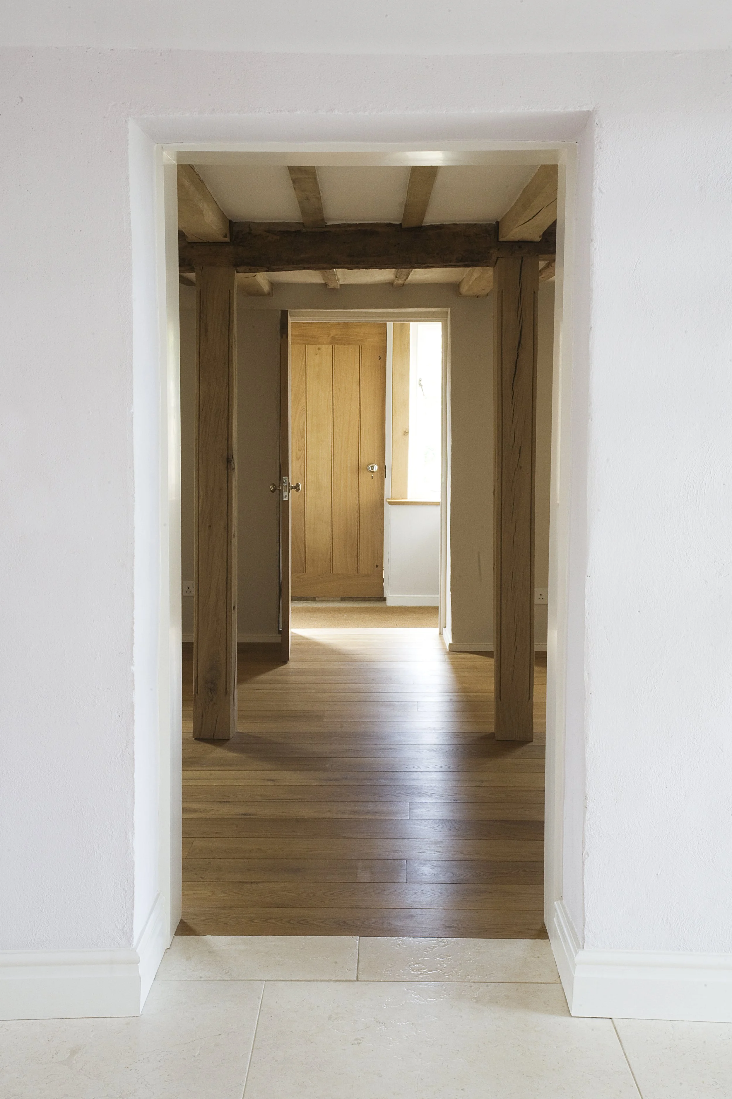 View through a doorway showing a room with wooden ceiling beams, a wooden door, and a window with natural light, with a space of tiled flooring outside the doorway.